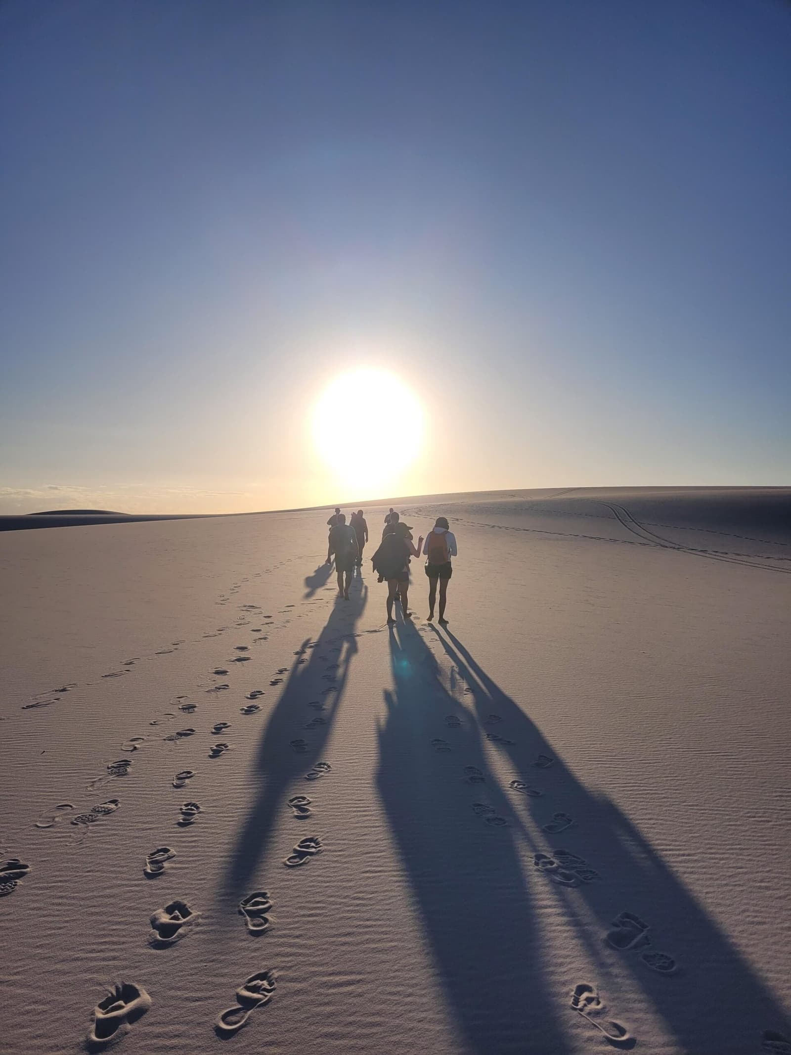 Long shadows, bare feet, and nowhere else to be. A small group of trekkers walks toward a low sun across wide white sand dunes on the lencois maranhenses trek, their long shadows and barefoot prints stretching back across the smooth sand