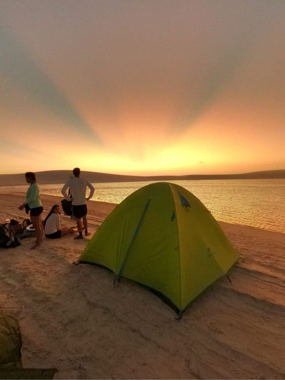 Your tent, a lagoon, and a sunset that earns its keep. A green dome tent pitched on a sandy riverbank during a Lençóis Maranhenses trekking expedition, with three travellers watching a vivid golden sunset over calm water and distant dunes