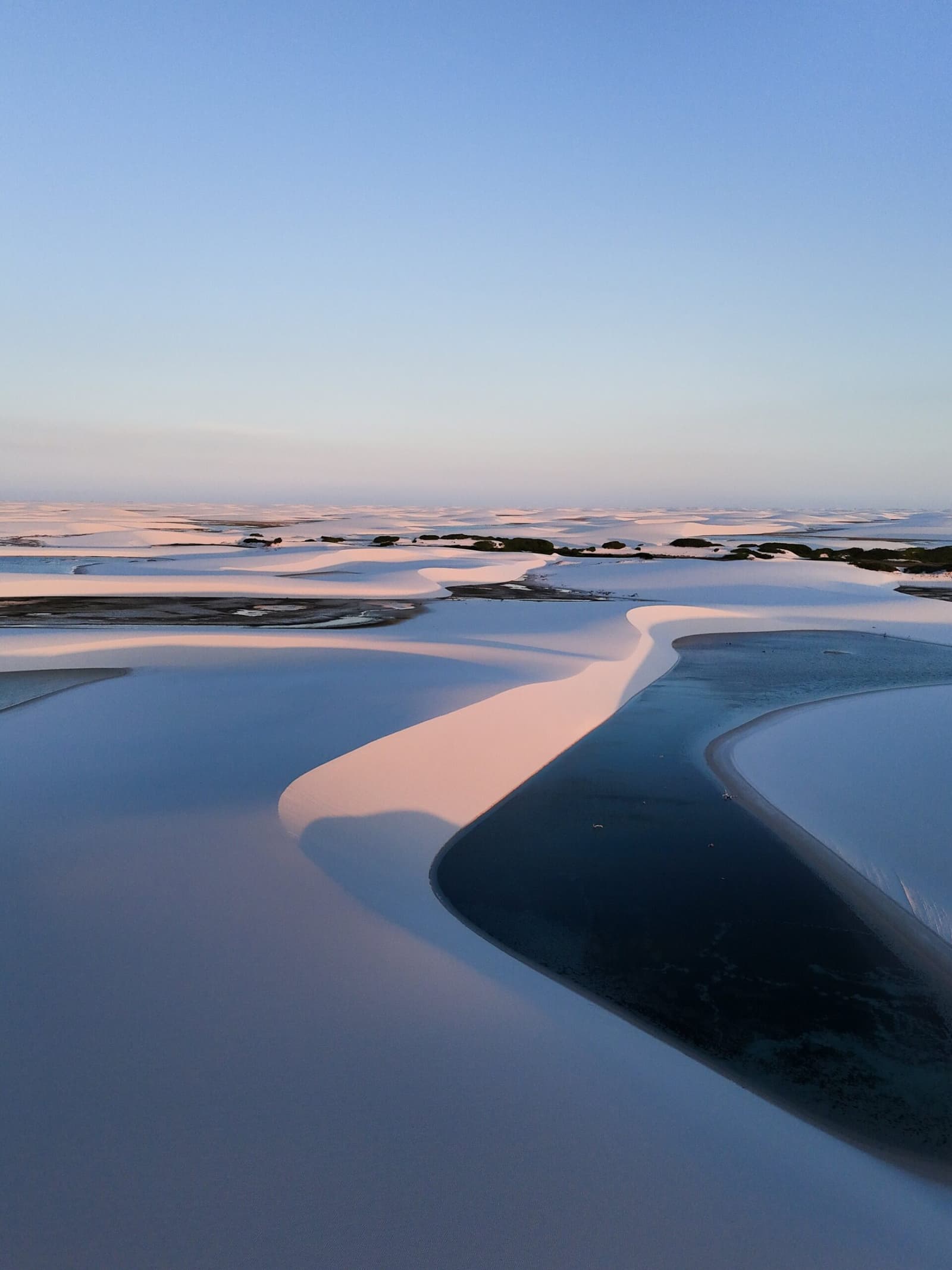 Pink light, cold water, and sand that goes on longer than you'd believe. Aerial view of white sand dunes and dark blue lagoons at dusk during a Lençóis Maranhenses trekking route, with warm pink light catching the dune ridges and a pale blue sky at the horizon