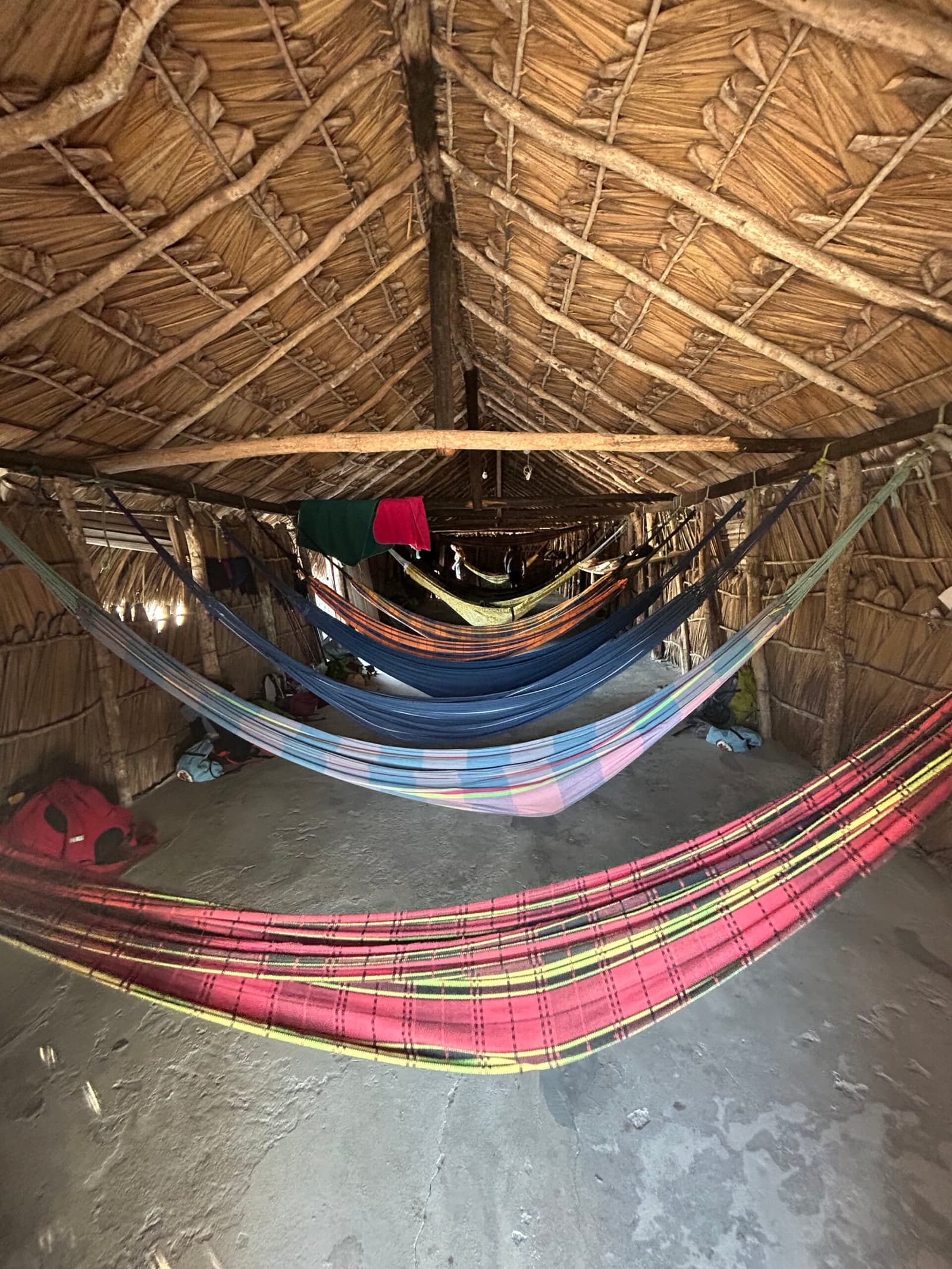 Your bed for the night: a hammock, a thatch roof, and no complaints. Colorful hammocks strung inside a traditional thatched-roof hut during a Lençóis Maranhenses trekking overnight stay, with traveler bags resting on the earthen floor