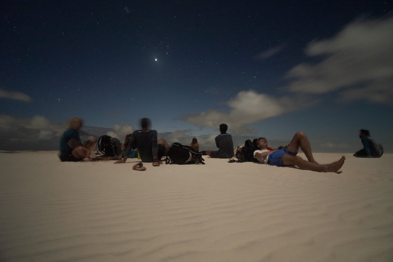 No chairs needed. Just sand, stars, and good company. A group of travelers lying and sitting on white sand dunes at night during a Lençóis Maranhenses trekking trip, gazing up at a star-filled sky with scattered clouds and a bright planet visible overhead