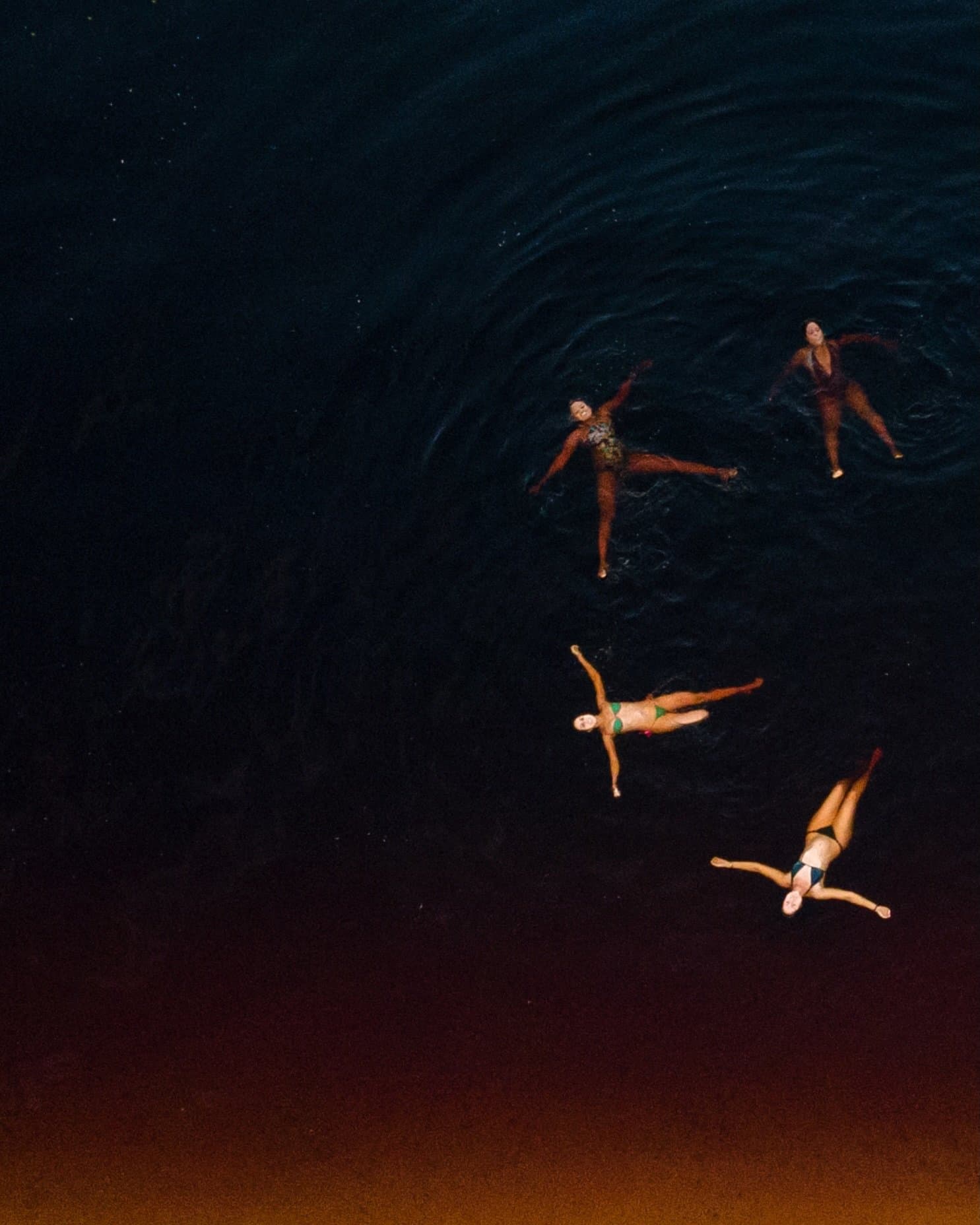 Four people, one dark river, zero hesitation. Aerial view of four swimmers floating spread-eagled on the dark black water of the Rio Negro near Mirante do Gavião, their colourful swimwear vivid against the deep indigo surface
