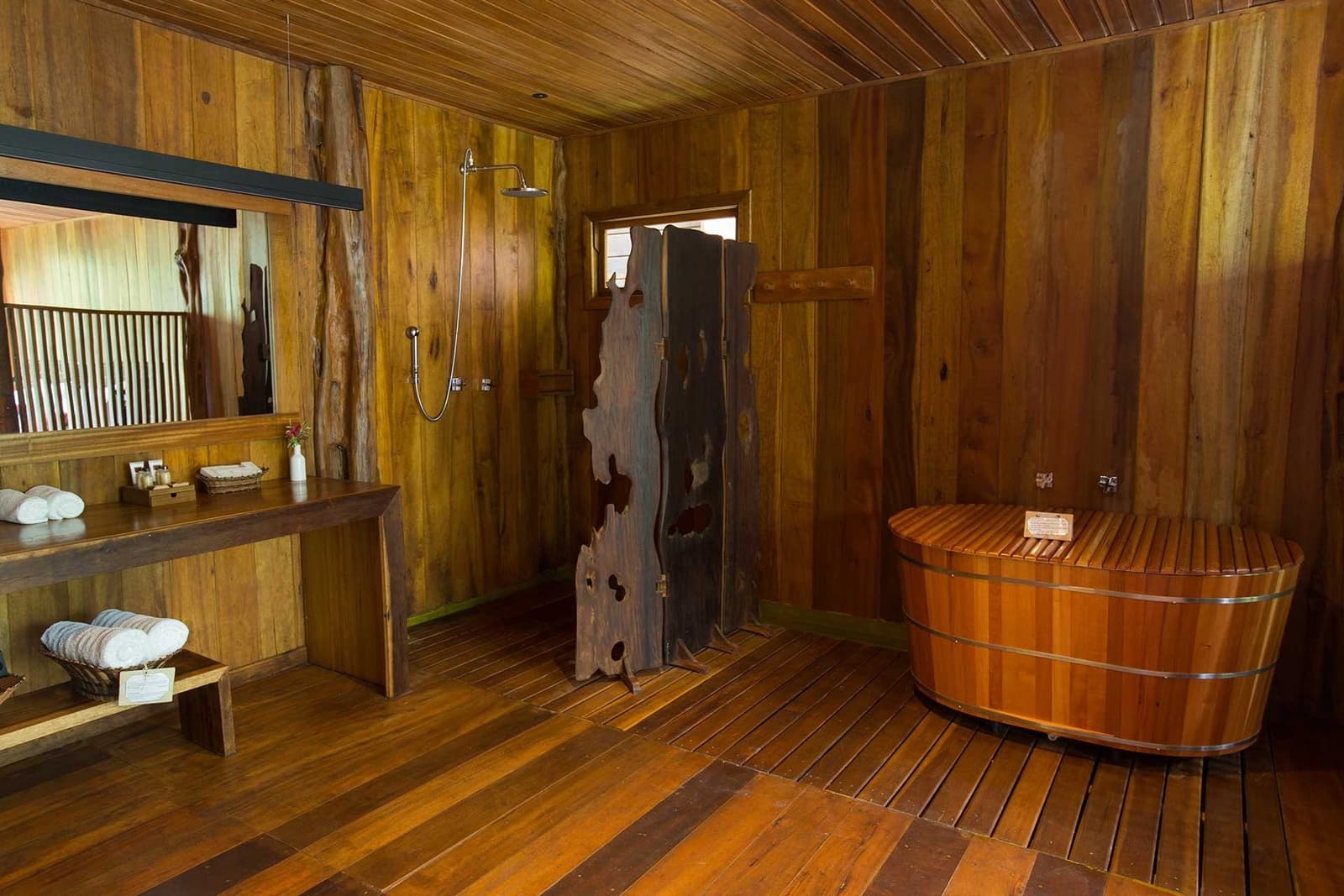 A hot soak in the forest, with the jungle just outside the window. Rustic all-wood bathroom at Mirante do Gavião featuring a barrel soaking tub, rainfall shower, live-edge timber divider, and folded white towels on a slatted bench