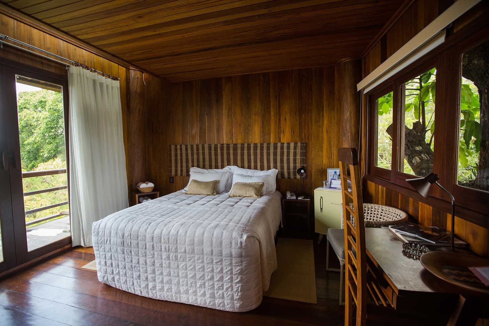 Sleep surrounded by forest, not separated from it. A Mirante do Gavião guest room with warm hardwood walls, ceiling, and floors, a white quilted double bed, woven headboard, wooden desk, and two windows overlooking lush green jungle foliage