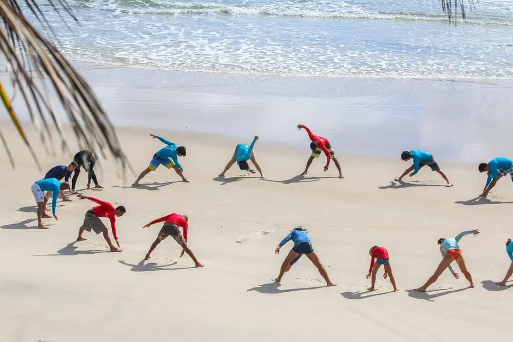 The warm-up circle that sorts the keen from the very keen. A group of students in rash guards performing stretching exercises on a white-sand beach before their surf lessons in Itacaré, with gentle ocean waves in the background and a palm frond in the foreground.
