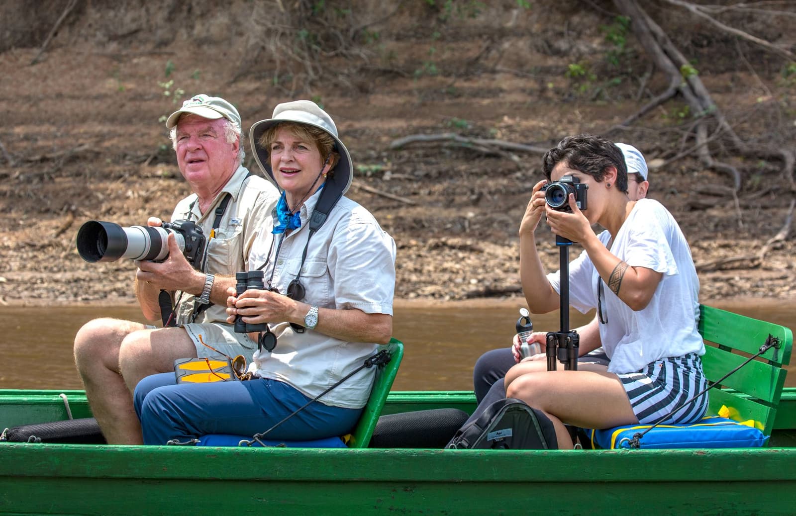 Cameras ready, river ahead, and something interesting just around the bend. Photo: Daniel Forster Three travelers on a green wooden boat along a river bank during a Tucano Amazon river cruise, one man holding a telephoto camera lens, a woman with binoculars, and a younger person shooting with a camera on a monopod