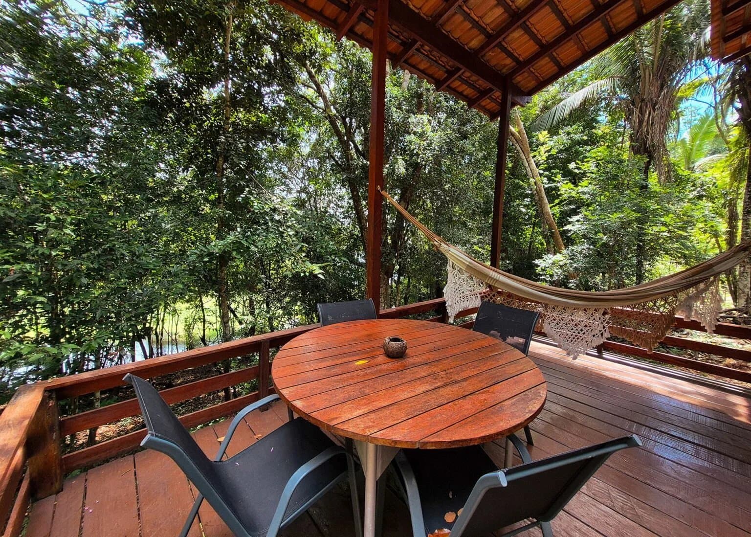 Lunch, a hammock, and the forest, pretty much the whole plan. Covered wooden deck at an Amazon boutique lodge with a round hardwood dining table, four chairs, two woven hammocks, and a lush rainforest canopy and waterway visible beyond the railing