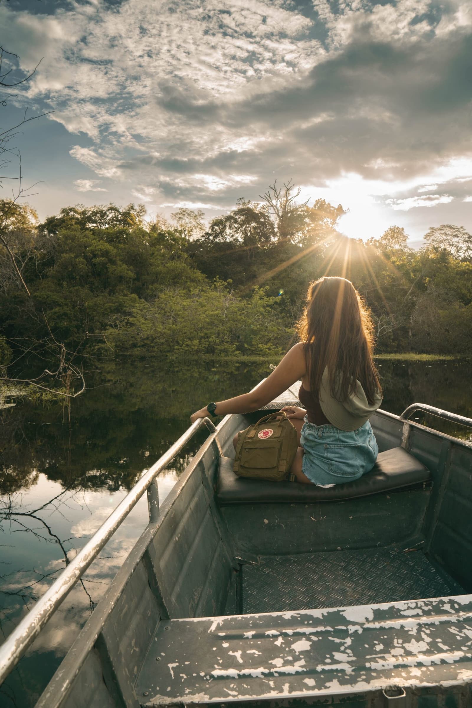 Some moments you don't photograph, you just sit there and let the river do its thing. Photo: Yuri Panichi A woman sits at the bow of a small aluminium boat on a calm river, watching the sun burst through the tree line at golden hour during an Amazon boutique lodge excursion