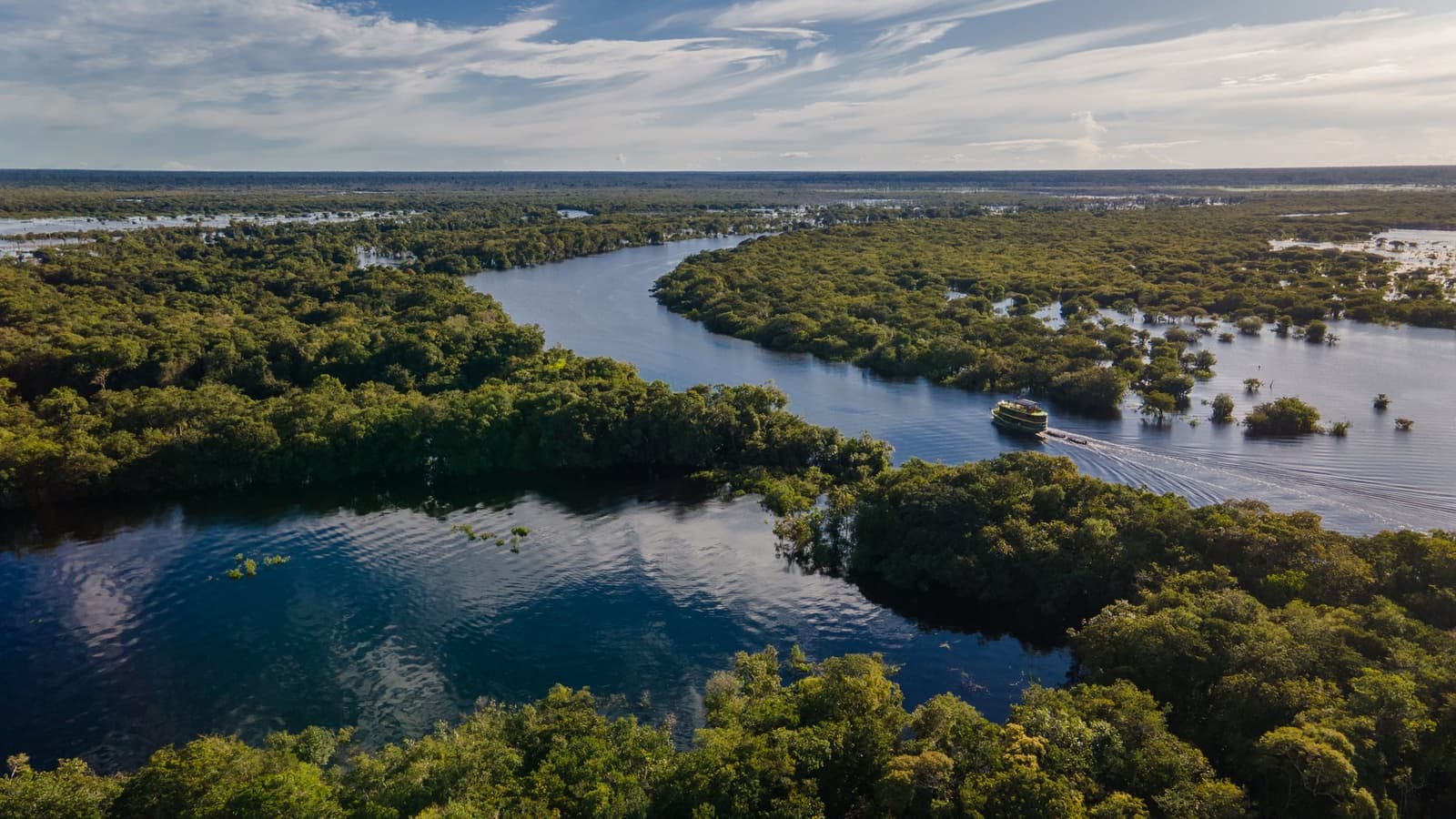 Out here, the river doesn't follow a map, it makes its own. Aerial view of a small cruise vessel navigating a winding Amazon river channel through flooded igapó forest on an Amazon Cruise Trip in Brazil, with vast flooded plains stretching to the horizon under a partly cloudy sky