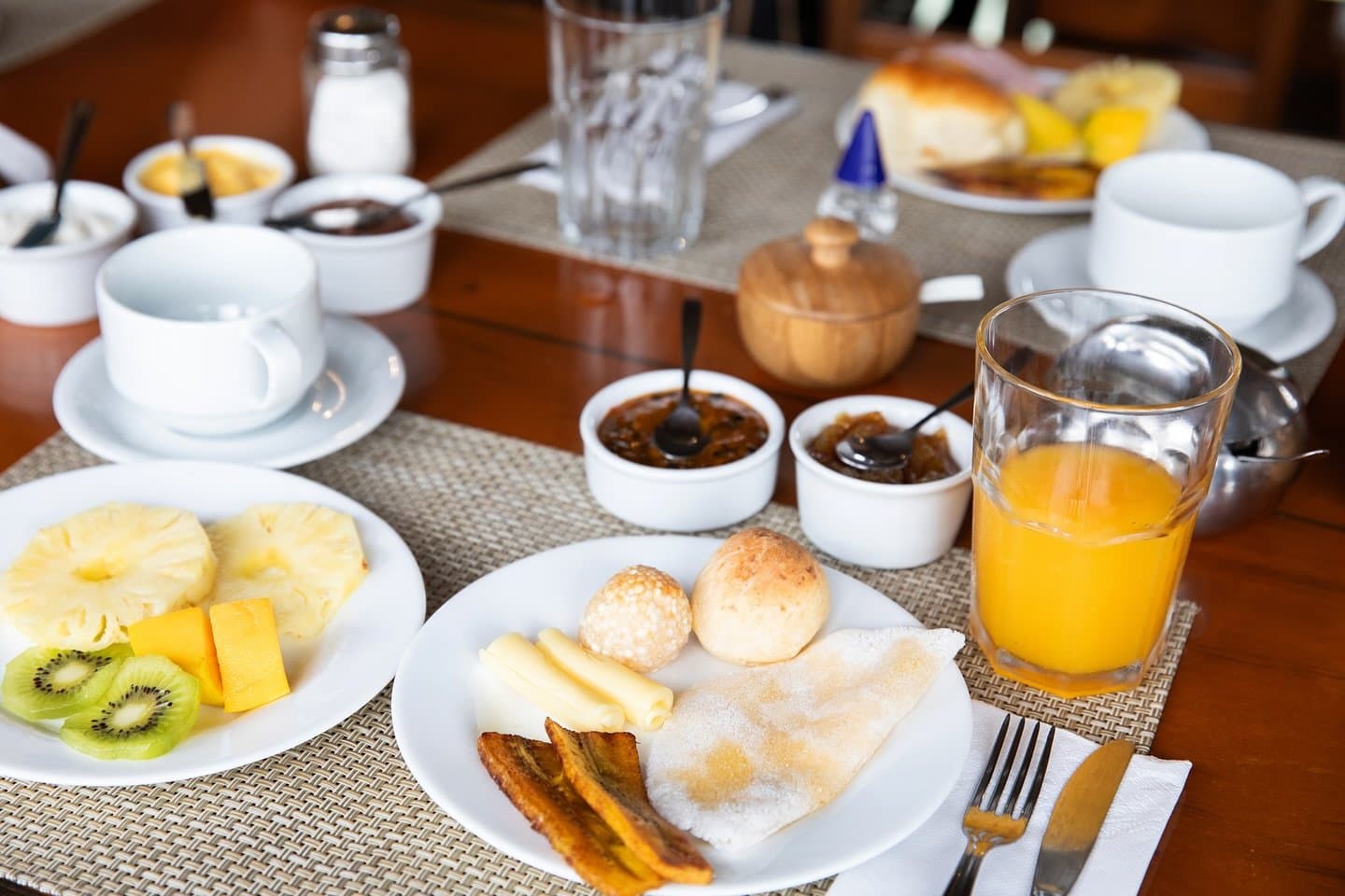 Fuel up right before the river calls you back. A full Amazon jungle cruise Brazil breakfast table set with fresh pineapple, kiwi and mango, cheese rolls, fried plantains, a tapioca crepe, fruit preserves and a glass of orange juice