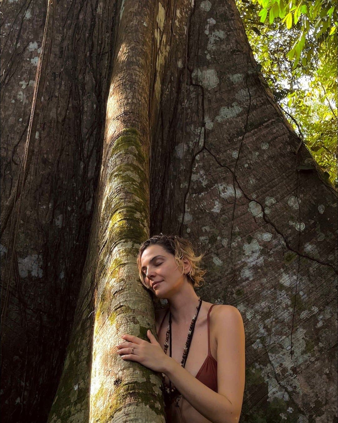 Some trees just ask to be hugged, and this one has clearly been asked before. A woman rests her cheek against the massive moss-covered trunk of a century-old rainforest tree during an amazon river cruise brazil itinerary, eyes closed, arms wrapped around it, with towering buttress roots rising behind her into the forest canopy