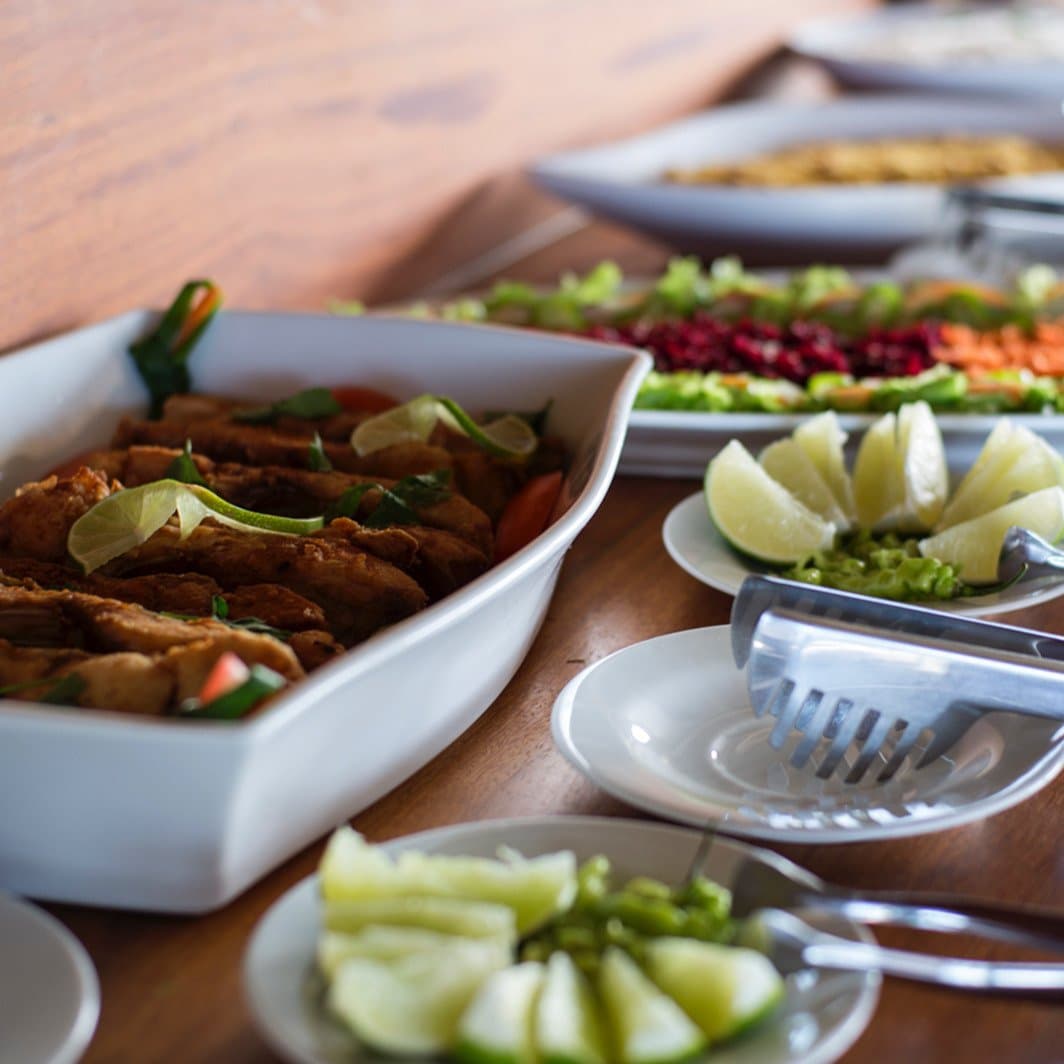 Good food mid-river: proof the Amazon asks nothing of your appetite. A buffet spread on a wooden table during an amazon river cruise brazil meal service, featuring a tray of golden fried fish fillets garnished with lime wedges and herbs, a colorful salad platter with beets, carrots and greens, and small side dishes of sliced cucumber and lime