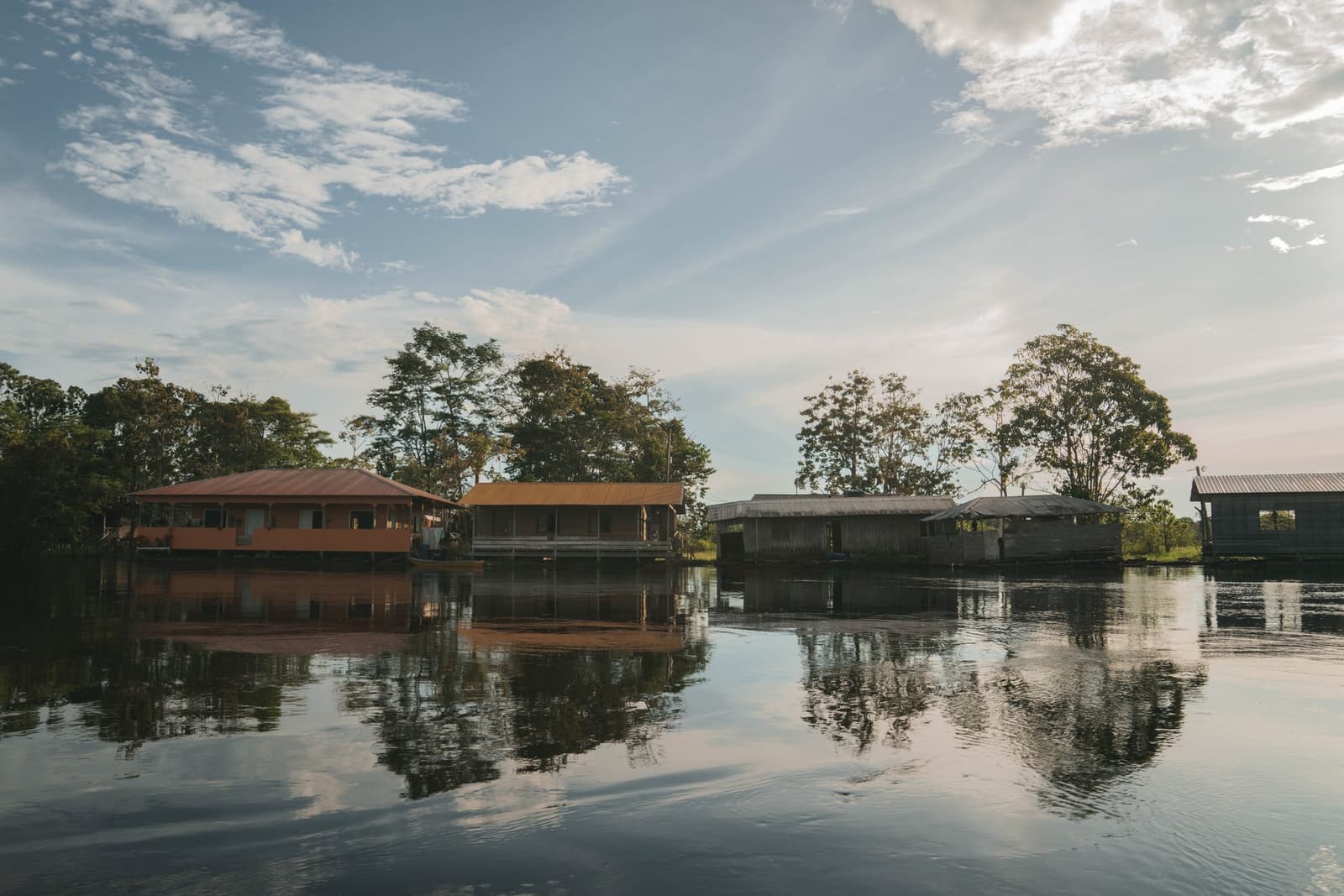 Your home for the week, mirrored perfectly in the river below. Stilted wooden and painted buildings of the Amazon Turtle Lodge sitting directly over a calm, dark river, with tall rainforest trees reflected in the still water beneath a partly cloudy sky