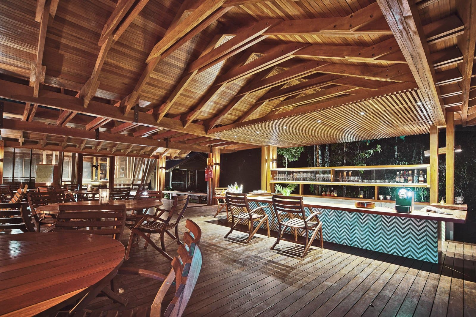 Cold drink sorted, forest humming just past the railing. Photo: Samuel Melim (c) Open-air bar and dining area at a birdwatching in the Amazon rainforest lodge, featuring exposed timber roof beams, a teal chevron-tiled bar counter lined with glassware and bottles, round wooden tables, and a view into the night forest beyond