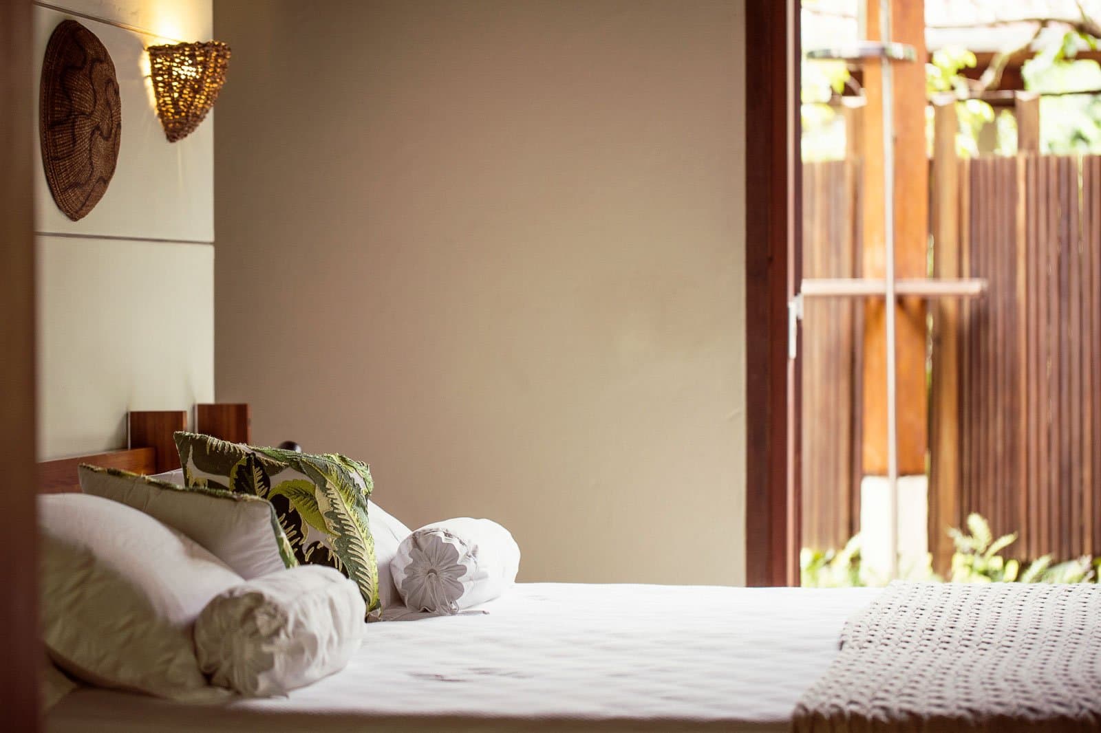 Rest up well, the birds start calling long before sunrise. Photo: | SamuelMelim Photography A neatly made double bed with tropical-print and white pillows in a birdwatching Amazon rainforest lodge room, with a woven wall sconce, handcrafted décor, and an open sliding door revealing a wooden deck and garden greenery