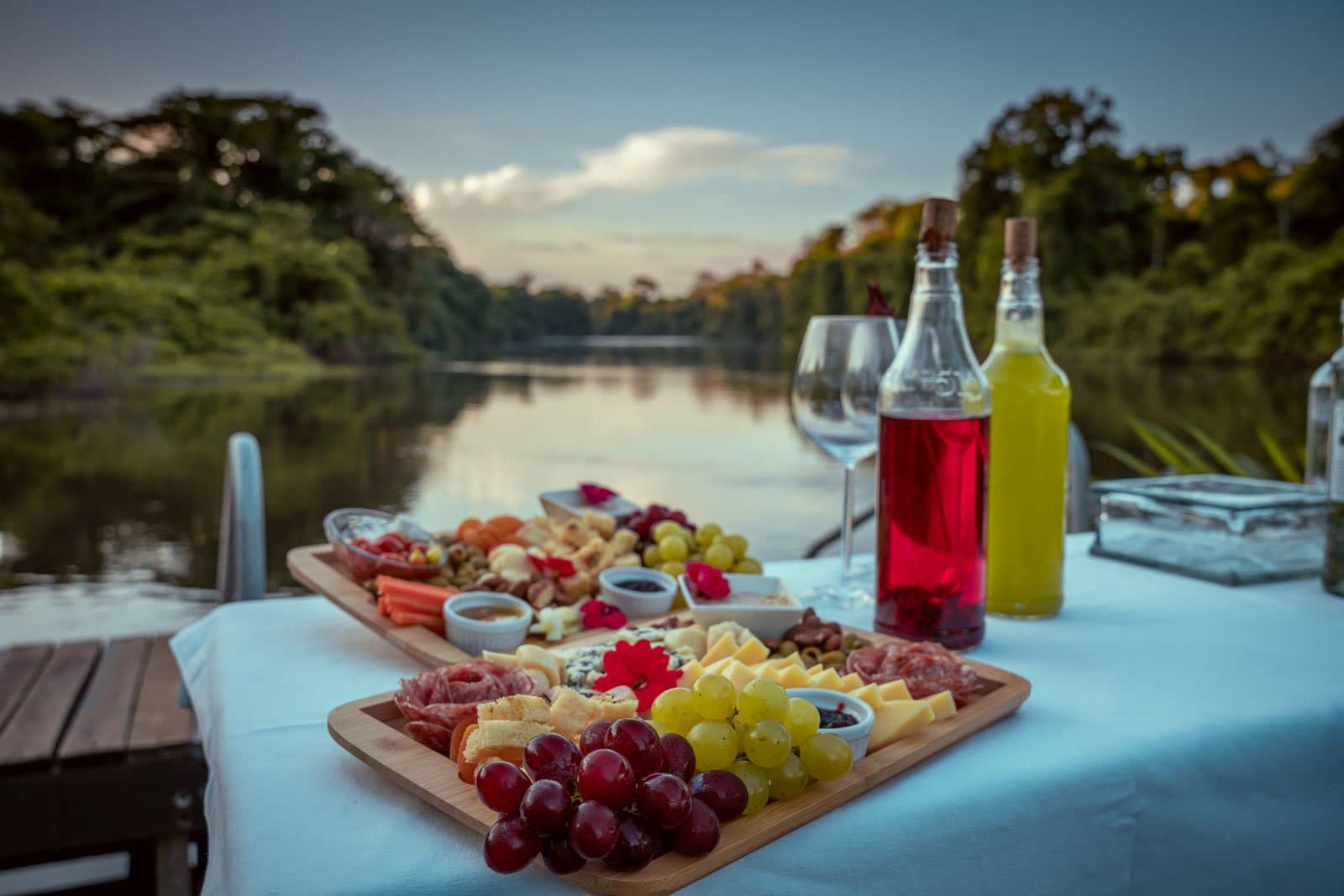 The river holds still, the board does not disappoint, and the forest can wait five more minutes. Photo: Samuel Melim A generous charcuterie board with grapes, cured meats, cheeses, and dipping sauces set on a white-clothed table beside a calm jungle river at dusk, with two bottles of colourful drinks and wine glasses, part of the birdwatching in the Amazon rainforest experience at Cristalino Jungle Lodge