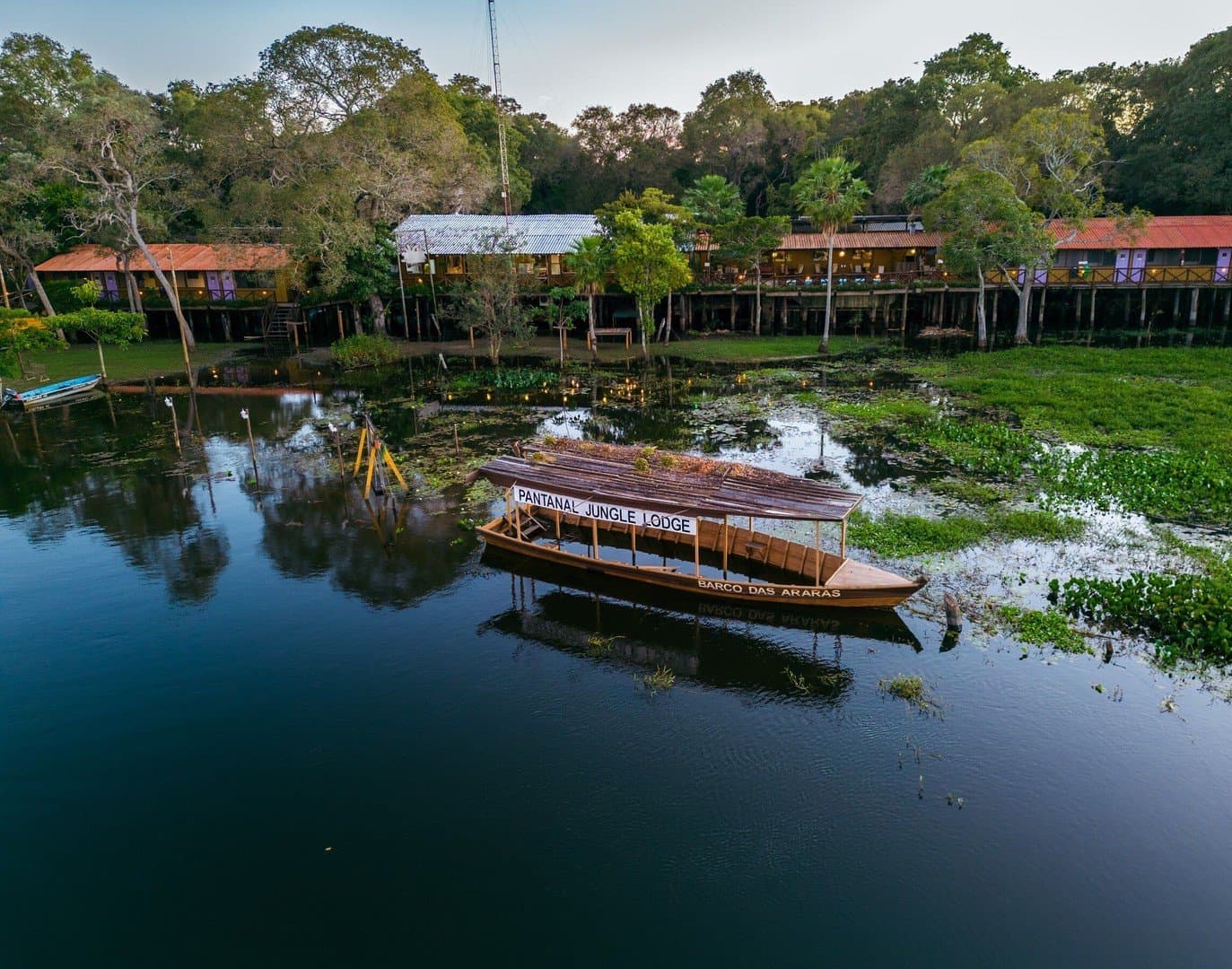 Your base camp floats in, quite literally. Aerial view of the Pantanal Jungle Lodge on a calm waterway, with the named wooden tour boat 'Barco das Araras' moored in the foreground among aquatic vegetation, and stilted lodge buildings nestled in dense trees on the bank — the base for this budget Pantanal tour