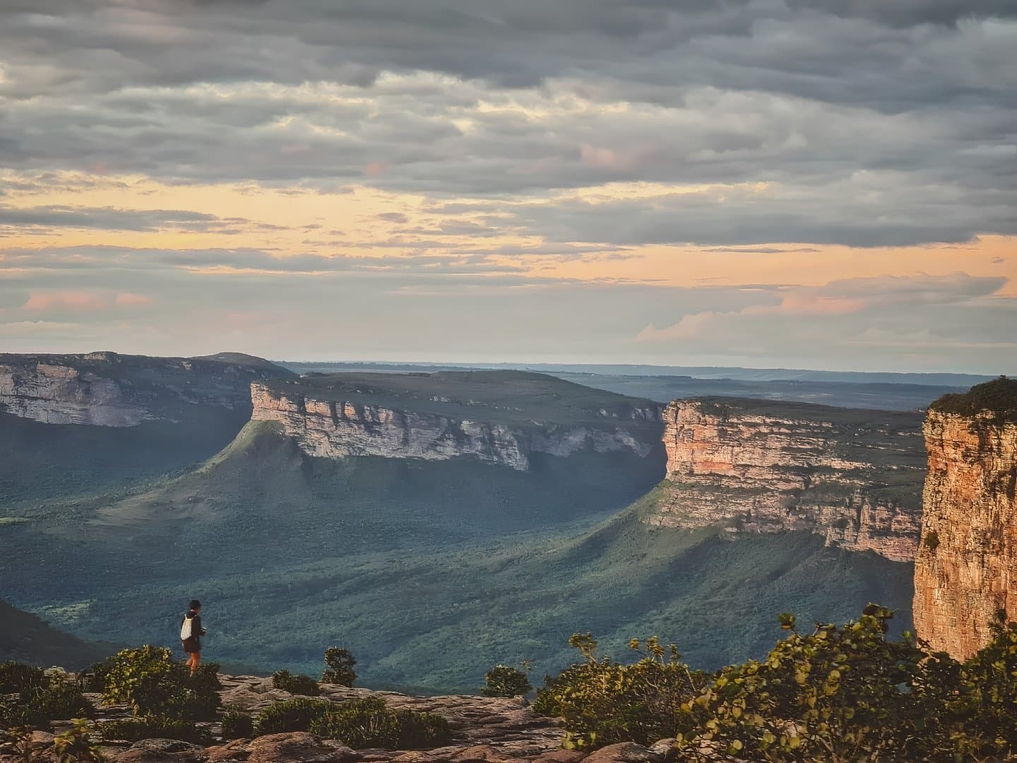 That moment when the canyon reminds you how small your to-do list really is. A lone hiker stands on a rocky ledge at sunset during a Chapada Diamantina Tour, gazing over the vast green valley and sandstone tabletop cliffs of Morro do Pai Inácio under a dramatic cloudy sky