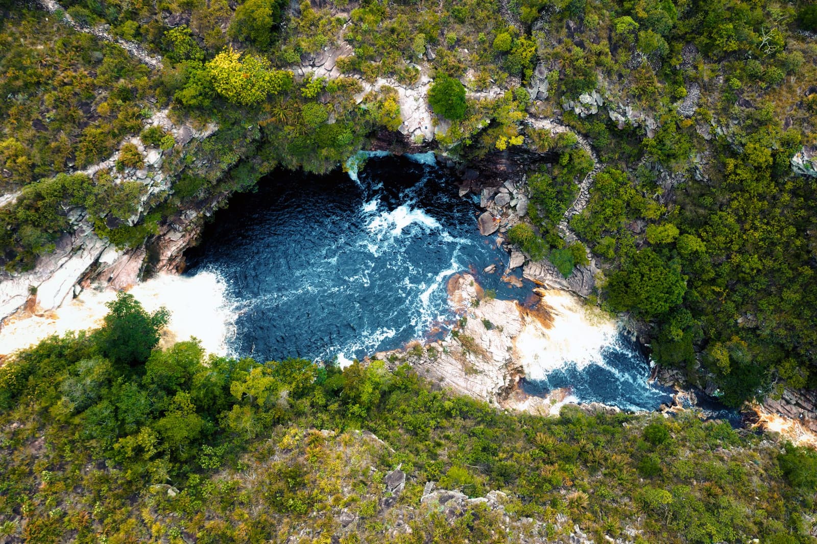 From above, the canyon keeps its best secret close to its chest. Aerial view of a deep blue natural pool surrounded by rocky outcrops and dense cerrado scrub vegetation, seen on a Chapada Diamantina Tour