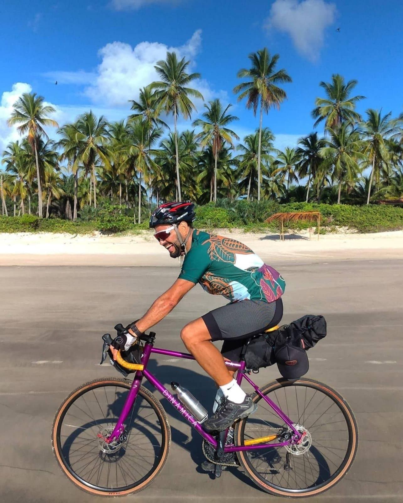 That grin says the road is flat and the sea breeze is on your side. A smiling cyclist on a loaded purple gravel bike rides a coastal road lined with tall coconut palms and white sand beach under a bright blue sky, capturing the spirit of cycling Bahia's scenic southern coast.