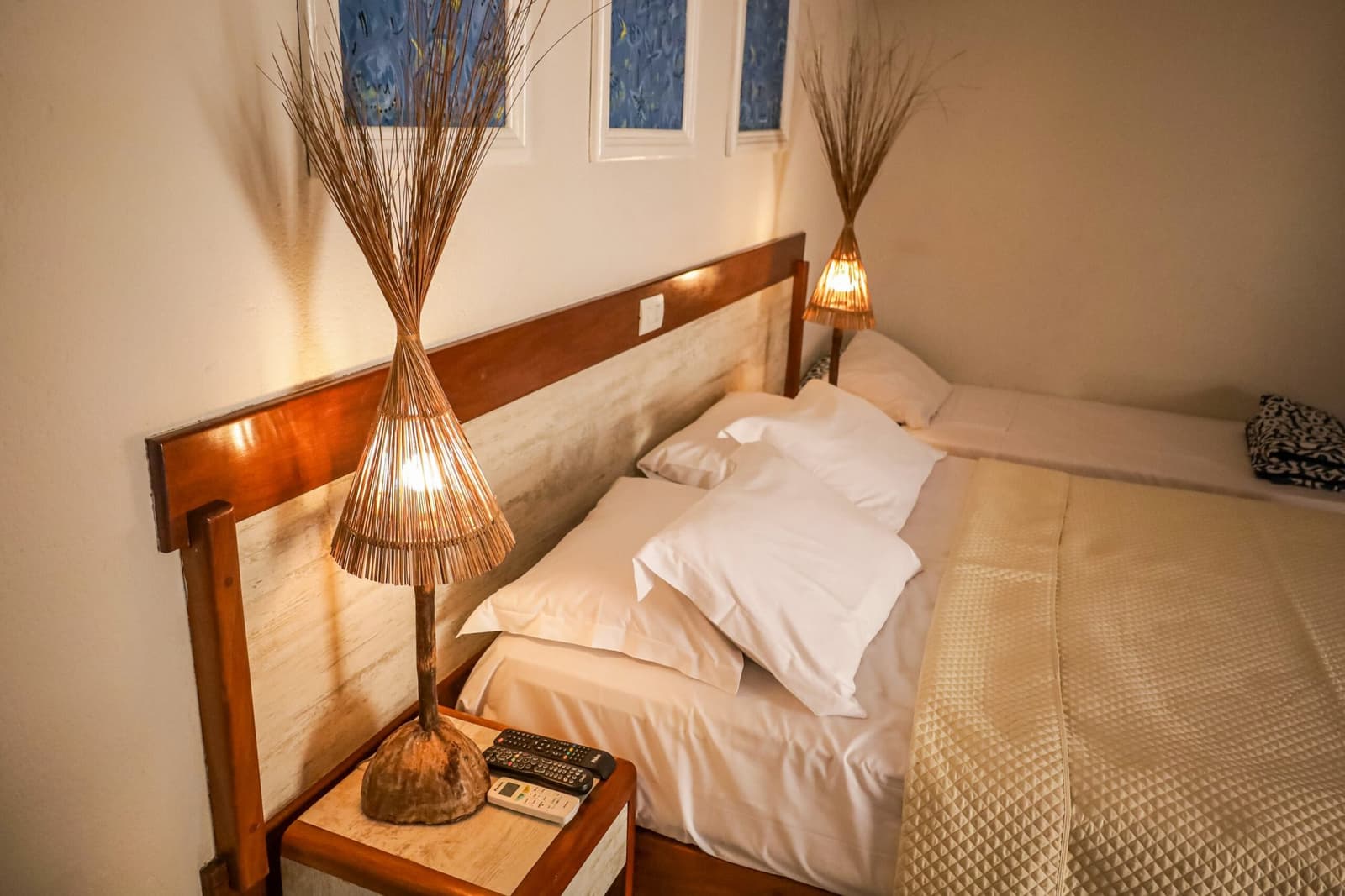 Small room, good sleep, the kind of night that makes tomorrow's ride feel easy. A warm, simply furnished guesthouse room on the cycling Bahia Cocoa Route, with two beds dressed in white linens, handcrafted rattan lamps on a wooden nightstand, and blue decorative windows above the headboard
