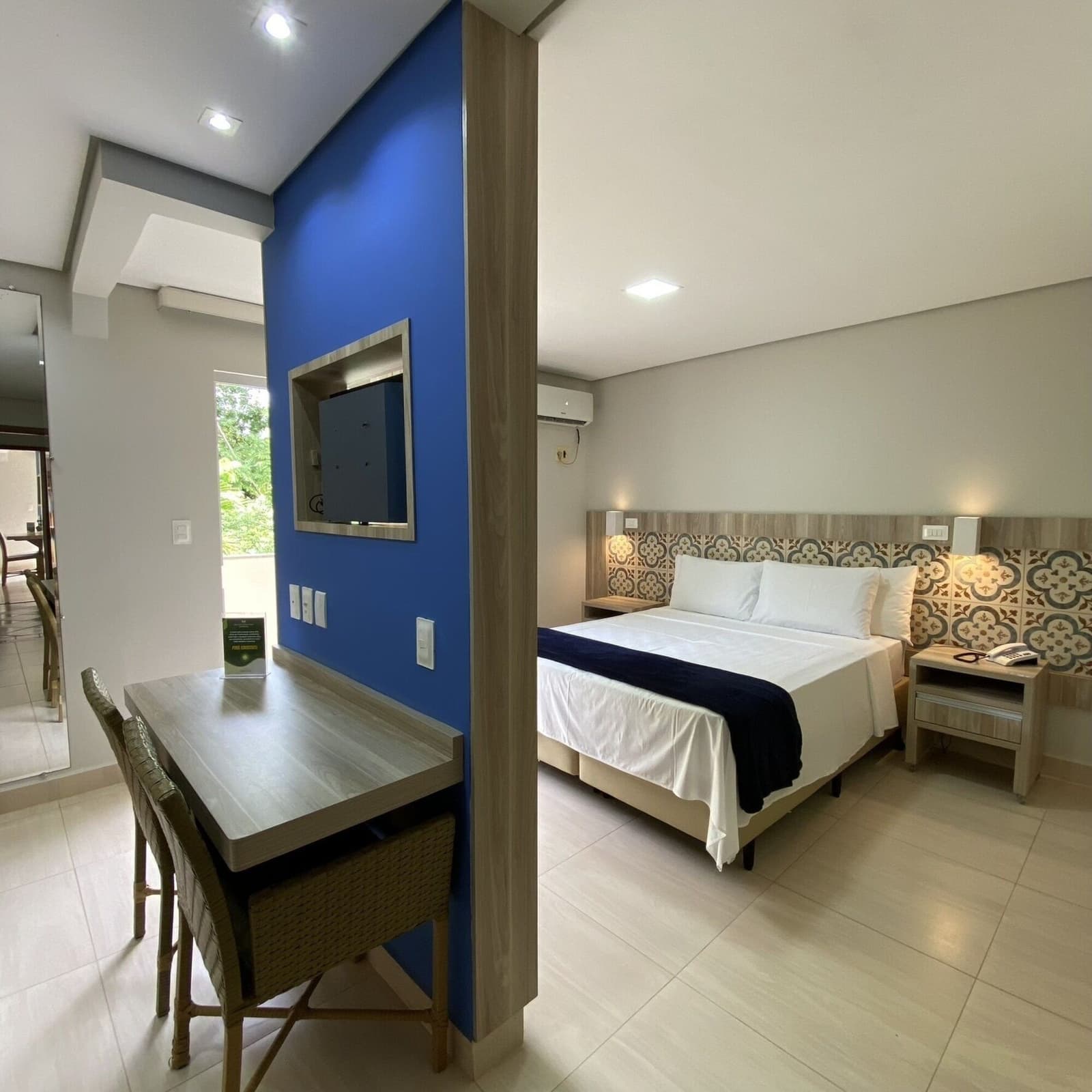 Good legs need a good night's sleep. This one delivers. Clean hotel room with a double bed, patterned headboard tiles, wooden desk, rattan chairs, and a bold blue accent wall with a wall-mounted TV, prepared for cycling bahia guests