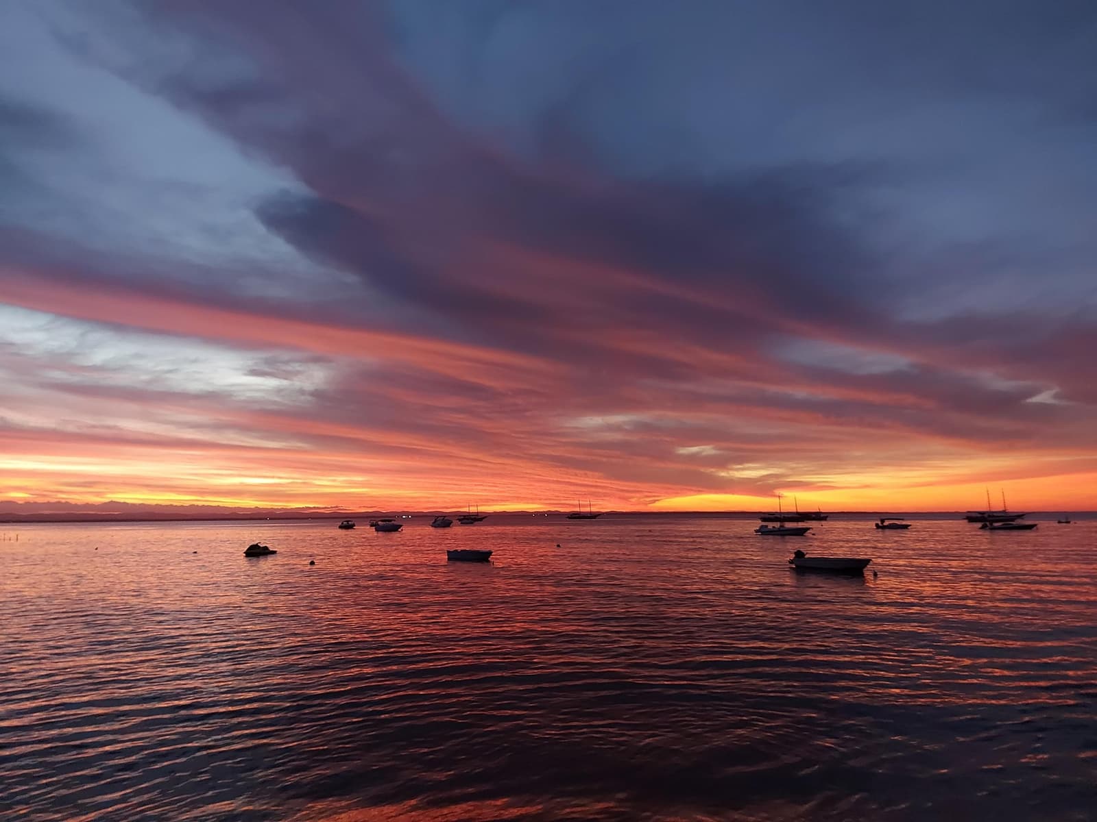 The bay earns its keep at dusk, no pedalling required. Fiery red and orange sunset over a calm bay with silhouetted fishing boats at anchor, seen from the Itacaré waterfront on the cycling Bahia cocoa route
