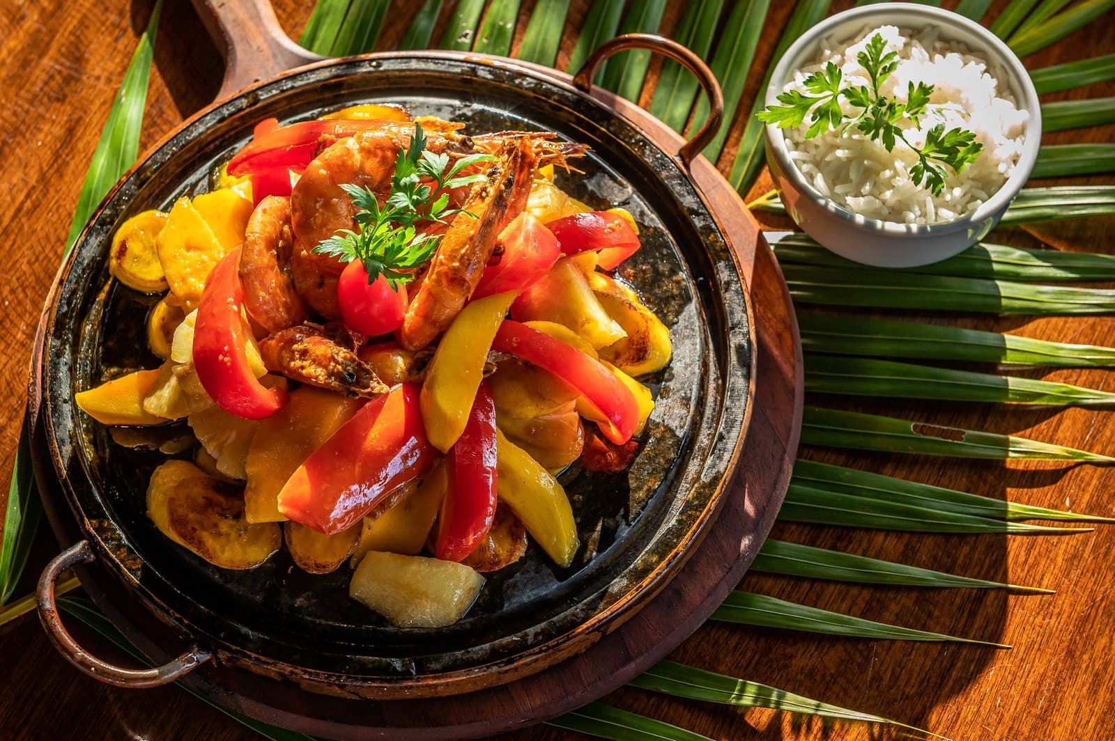 Forty kilometres of coastline, then this arrives at the table. No complaints. Sizzling clay skillet of whole grilled shrimp with red and yellow bell peppers, served on a wooden board with a side of white rice garnished with parsley, set on palm fronds — the kind of fuel you earn on a cycling Bahia tour