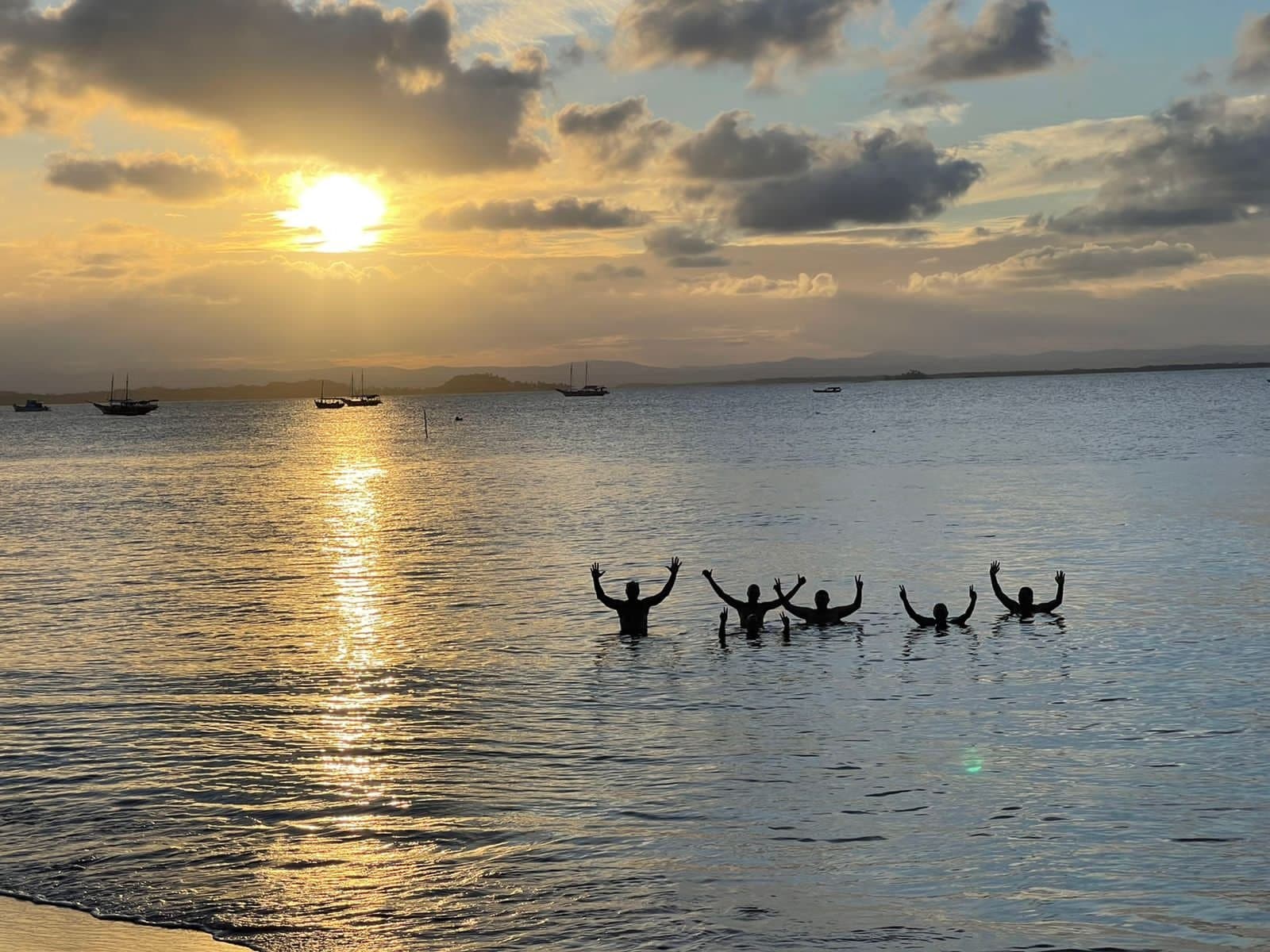 Arms up, sun down, the Rio das Contas delivers its verdict on the day. Five people silhouetted in the water with arms raised, celebrating a golden cycling Bahia sunset over a calm bay with sailboats anchored in the distance
