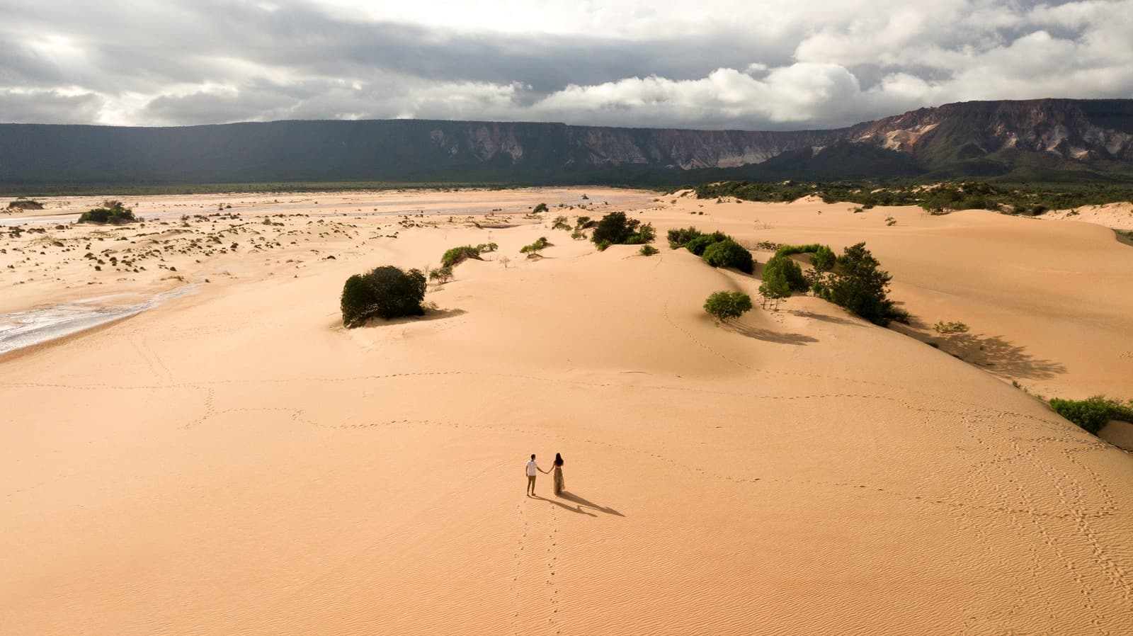Two people, a sea of sand, and footprints going exactly where they want. Photo: FG Trade Aerial view of a couple holding hands on vast golden sand dunes during a Jalapão trekking journey, with green shrubs, a river, and a dramatic sandstone escarpment under a stormy sky