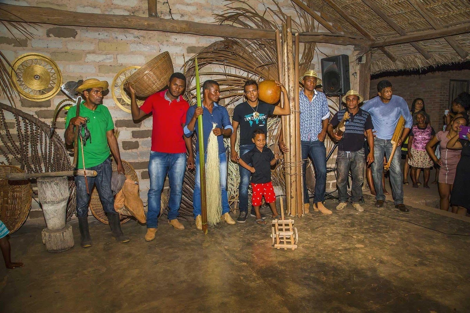 The Mumbuca community puts its whole story in your hands, one craft at a time. Local men and boys stand inside a thatched-roof brick building in the Mumbuca community, holding handcrafted objects including woven baskets, a gourd, bamboo poles and wooden tools as part of a cultural presentation during a Jalapão trekking visit