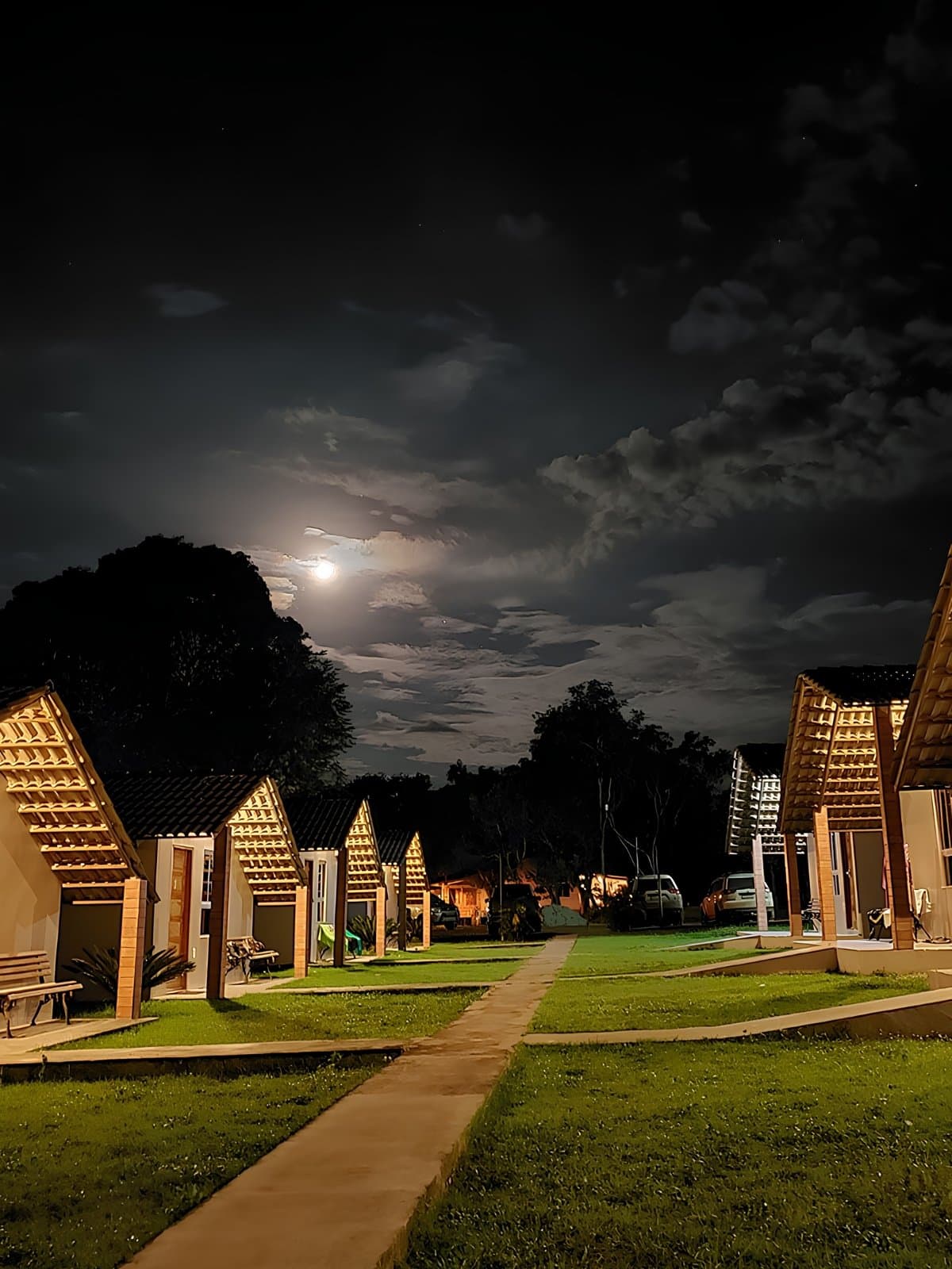 Quiet chalets, a full moon overhead, and tomorrow's trail already on your mind. A row of illuminated wooden-framed chalets with steep pitched roofs lines a grass pathway at night on a jalapão trip, with a full moon glowing through clouds above dark treetops
