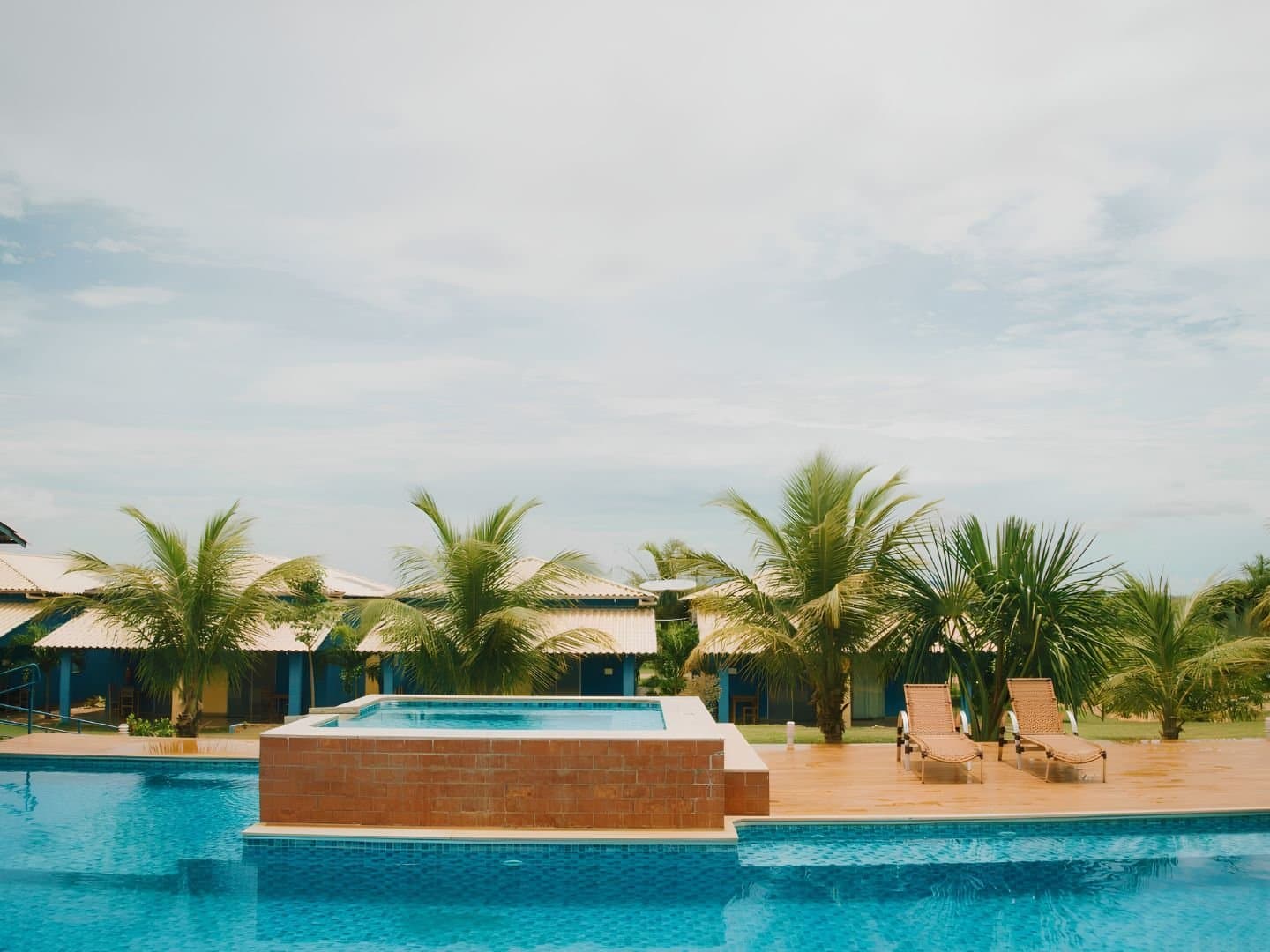 Cold pool, warm evening, no phone signal. That's the plan. Outdoor swimming pool with a raised jacuzzi and two sun loungers on a tiled deck, palm trees and blue-painted guesthouse bungalows in the background at a jalapão trip lodge