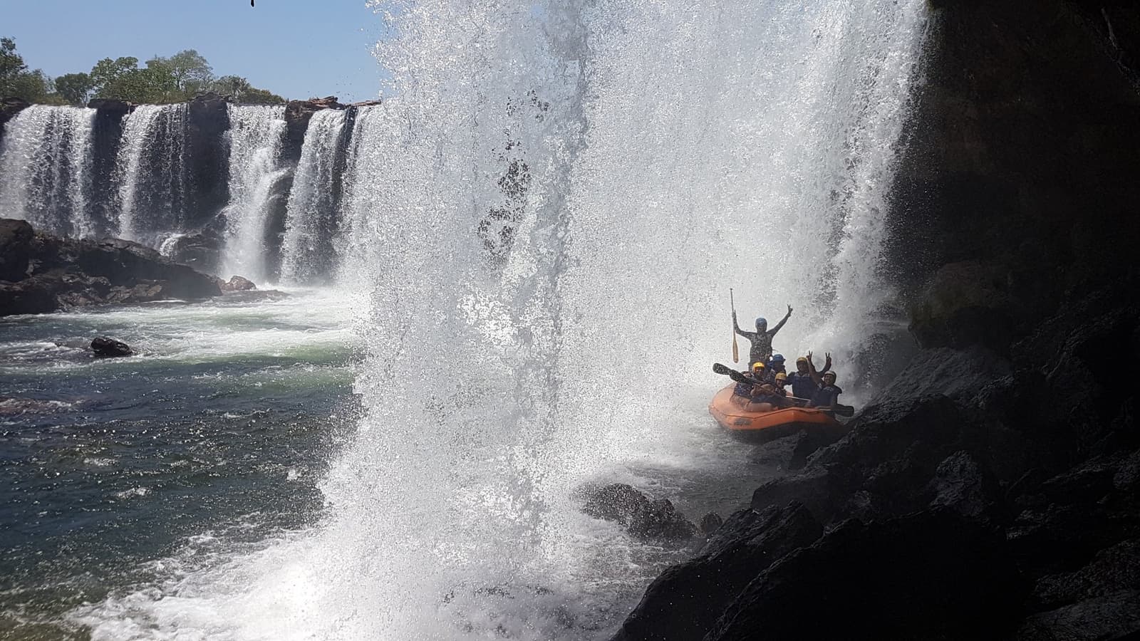Getting soaked was never a question. Only how soaked. A group of helmeted rafters in an orange inflatable raft sheltering beside dark rocks as a massive waterfall curtain crashes down during a jalapão trip on the Soninho River