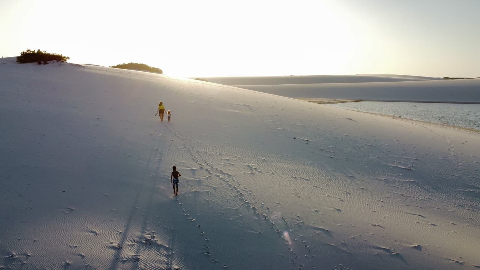 The dune doesn't care how small you are, it waits at the top either way. An adult and two children climb a vast white sand dune beside a still lagoon during the golden hour on a Lençóis Maranhenses full itinerary