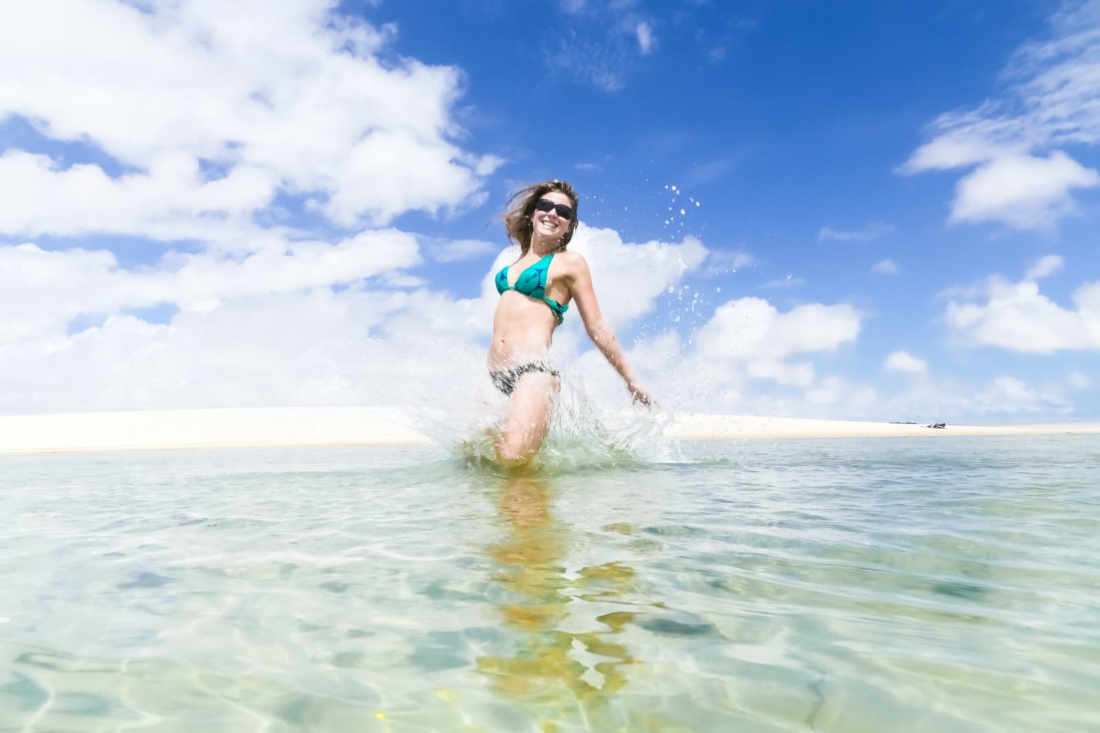 Cold water, warm sand, zero regrets. A smiling woman in a teal bikini splashes through crystal-clear shallow lagoon water against white sand dunes and a vivid blue sky in Lençóis Maranhenses Full Itinerary