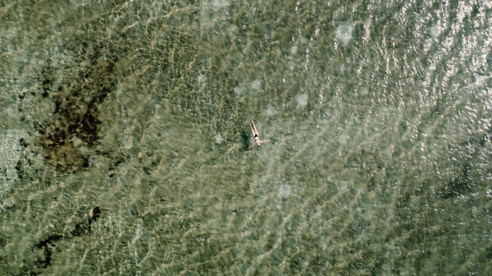 Just you, the water, and a drone that caught the whole thing. Aerial view of a lone swimmer floating on their back in shallow crystal-clear water over a patterned sandy and rocky seabed, captured from directly above as part of a Lençóis Maranhenses full itinerary