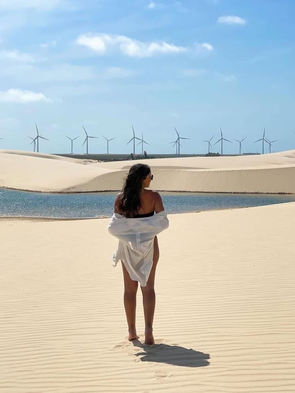 Sand, water, and spinning turbines, Lençóis Maranhenses does nothing by halves. A woman in a white shirt stands barefoot on rippled white sand dunes facing a sparkling blue lagoon, with a row of wind turbines stretching across the horizon under a bright sky — a striking scene from a Lençóis Maranhenses full itinerary
