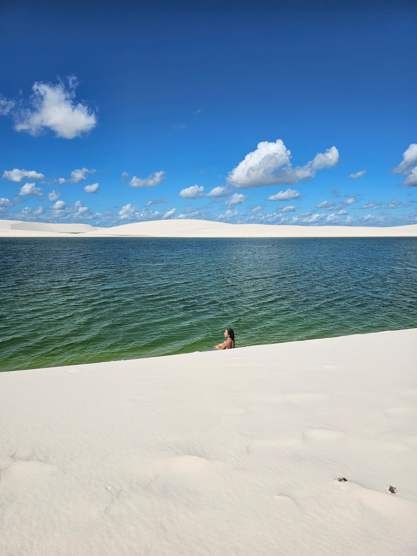 Just you, the green water, and a horizon made entirely of sand. A woman wading chest-deep in a clear green-blue freshwater lagoon on the Lençóis Maranhenses hike, with vast white dunes rising on the far shore beneath a bright blue sky
