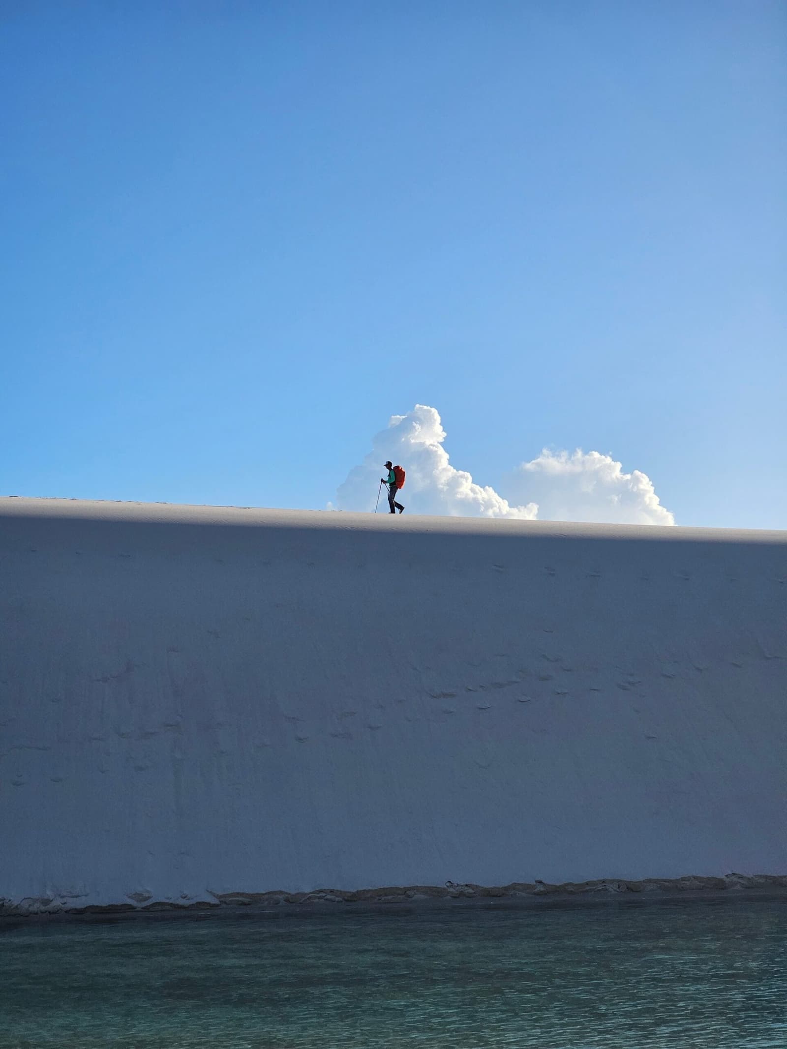 Small figure, big dune. That ridge puts everything in perspective. A lone hiker with a red backpack and trekking pole walks along the sharp ridge of a towering white sand dune during a Lençóis Maranhenses hike, with a turquoise lagoon at the base and billowing clouds behind