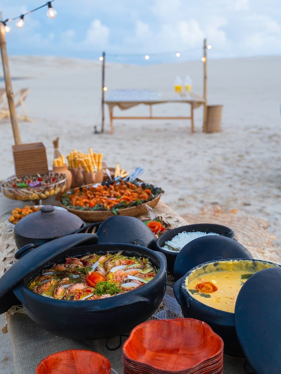 Dinner on the dunes: possibly the best place setting in all of Maranhão. A spread of traditional clay pot seafood dishes, including shrimp moqueca and a golden cream sauce, set on a rustic table on white sand dunes under string lights at dusk in Lençóis Maranhenses Santo Amaro