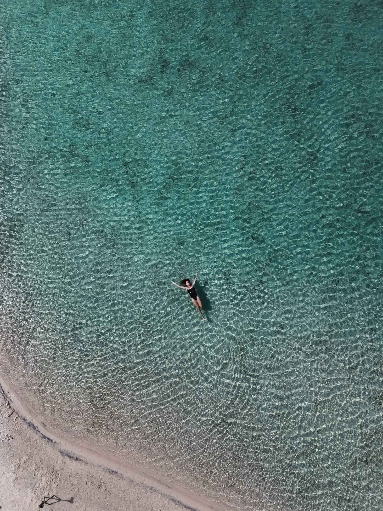 Just you, the water, and a drone you forgot was there. Aerial view of a woman floating on her back in shallow turquoise water near a white sand shore on the Lençóis Maranhenses trek from Atins