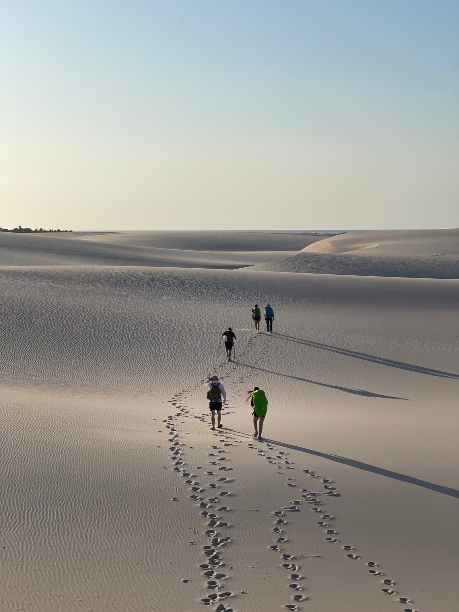 Four people, one enormous sandscape, and no wrong turns, just follow the footprints. Five hikers with backpacks walking across rippled white sand dunes on the Lençóis Maranhenses trek, their footprints trailing behind them under a pale early-morning sky