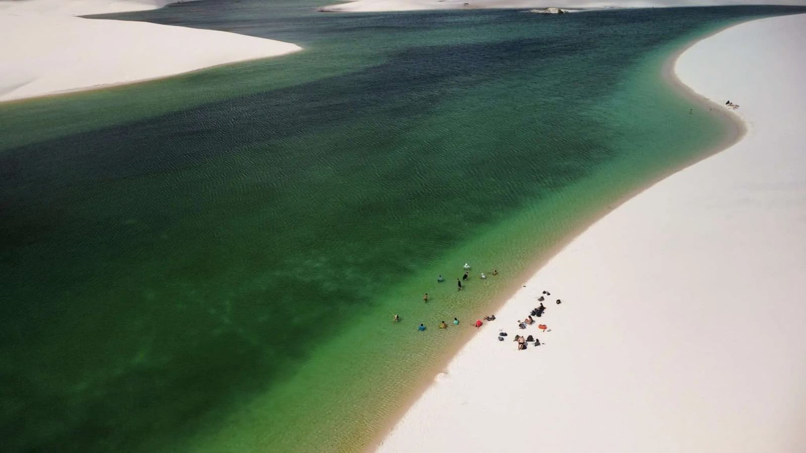 Tiny figures, enormous lagoon, scale has a way of putting things in perspective here. Aerial view of a large freshwater lagoon on the Lençóis Maranhenses trek, with deep green and teal water curving between bright white sand dunes and a small group of visitors wading at the shoreline