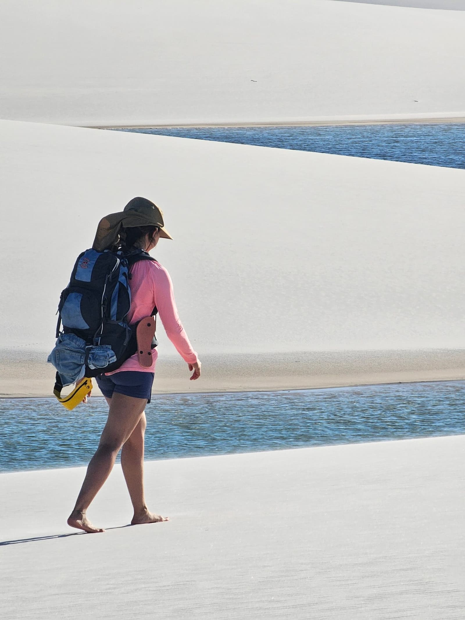 Shoes optional, sense of wonder required. A solo hiker with a backpack and sun hat walks barefoot across white sand dunes on the lencois maranhenses trek, with a clear blue lagoon cutting between the dune ridges in the background