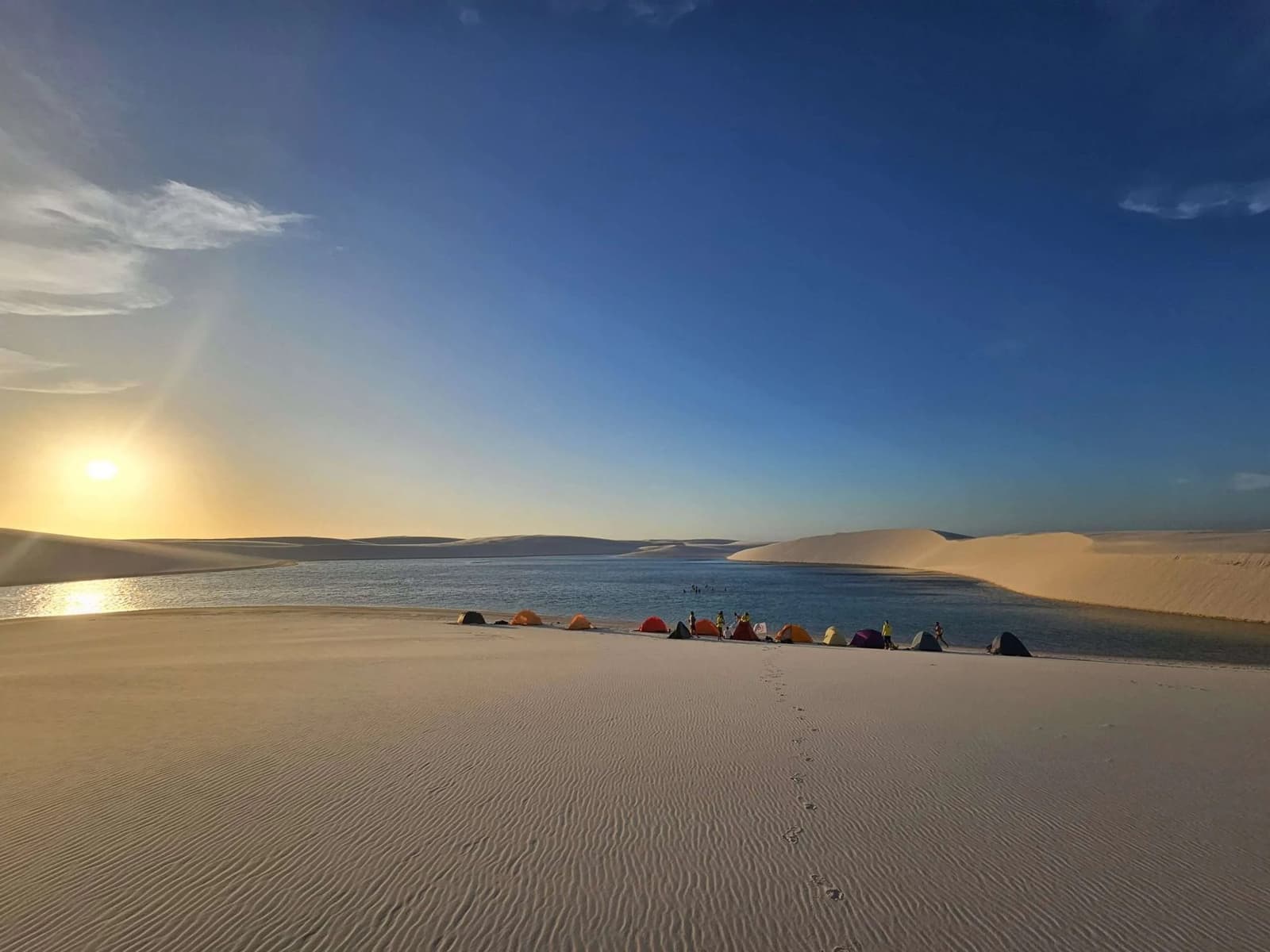 Your bedroom for the night: white dunes, a glassy lagoon, and no address. Colorful tents pitched along the shore of a freshwater lagoon at sunset during a Lençóis Maranhenses trekking expedition, with rippled white sand dunes stretching to the horizon under a deep blue sky