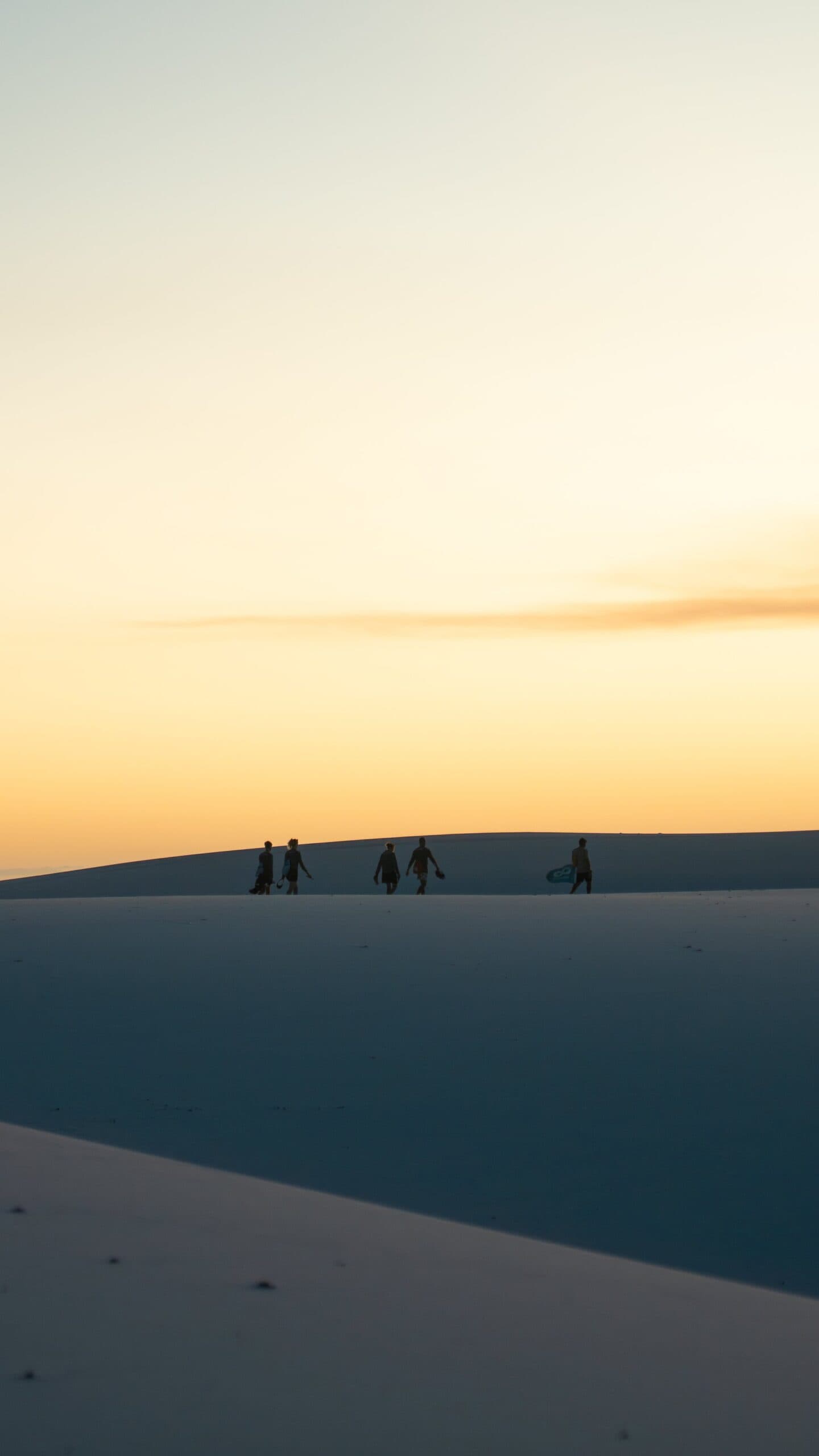 Last light, boards in hand, and a dune that feels like it goes on forever. Five silhouetted figures, one carrying a sandboard, walk along the crest of a white sand dune during a Lençóis Maranhenses trekking route at sunset, with a warm golden sky behind them