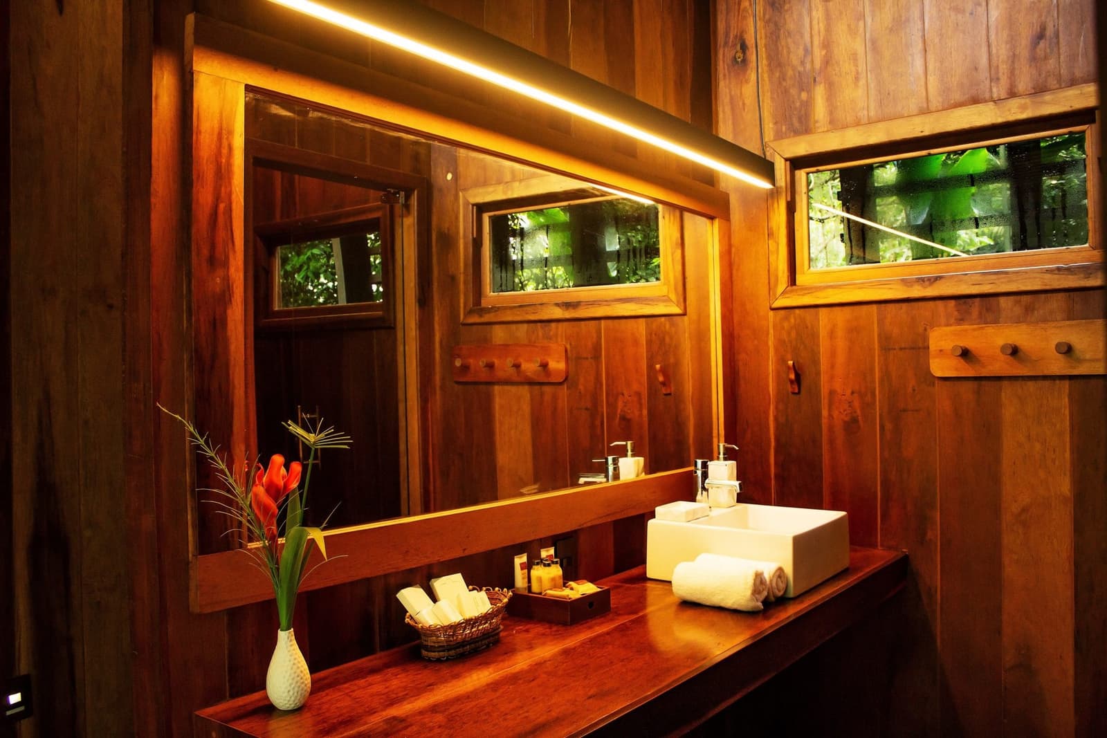 The forest is right outside, and somehow the bathroom knows it. Warm-lit bathroom at Mirante do Gavião featuring a polished hardwood vanity, vessel sink, fresh red tropical flowers, and jungle-view windows