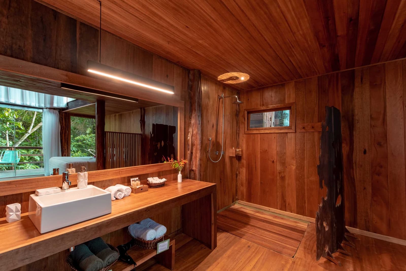 Rustic craftsmanship meets real comfort, the forest is right outside that window. Warm all-wood bathroom at Mirante do Gavião with a vessel sink vanity, rainfall shower, folded white towels, and a jungle view through the window