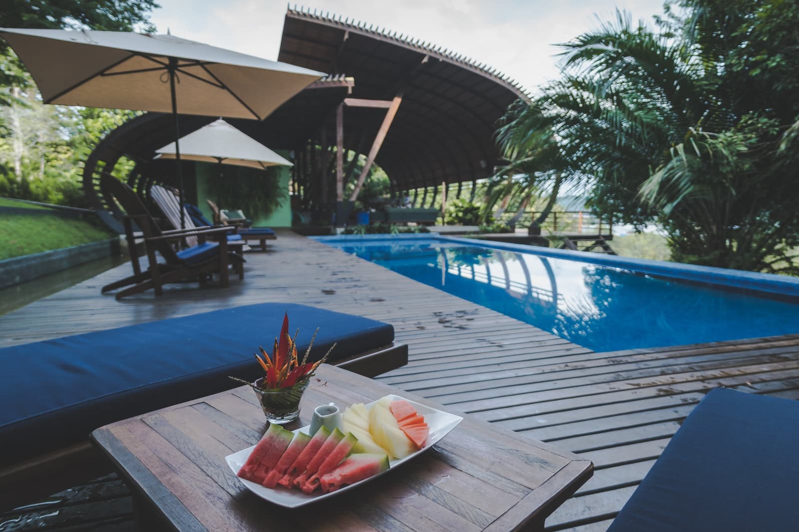 Fresh fruit, a cool pool, and the forest right there. Not a bad afternoon. A plate of sliced watermelon and pineapple on a poolside lounger table at Mirante do Gavião, with a curved-roof open pavilion and lush tropical greenery in the background